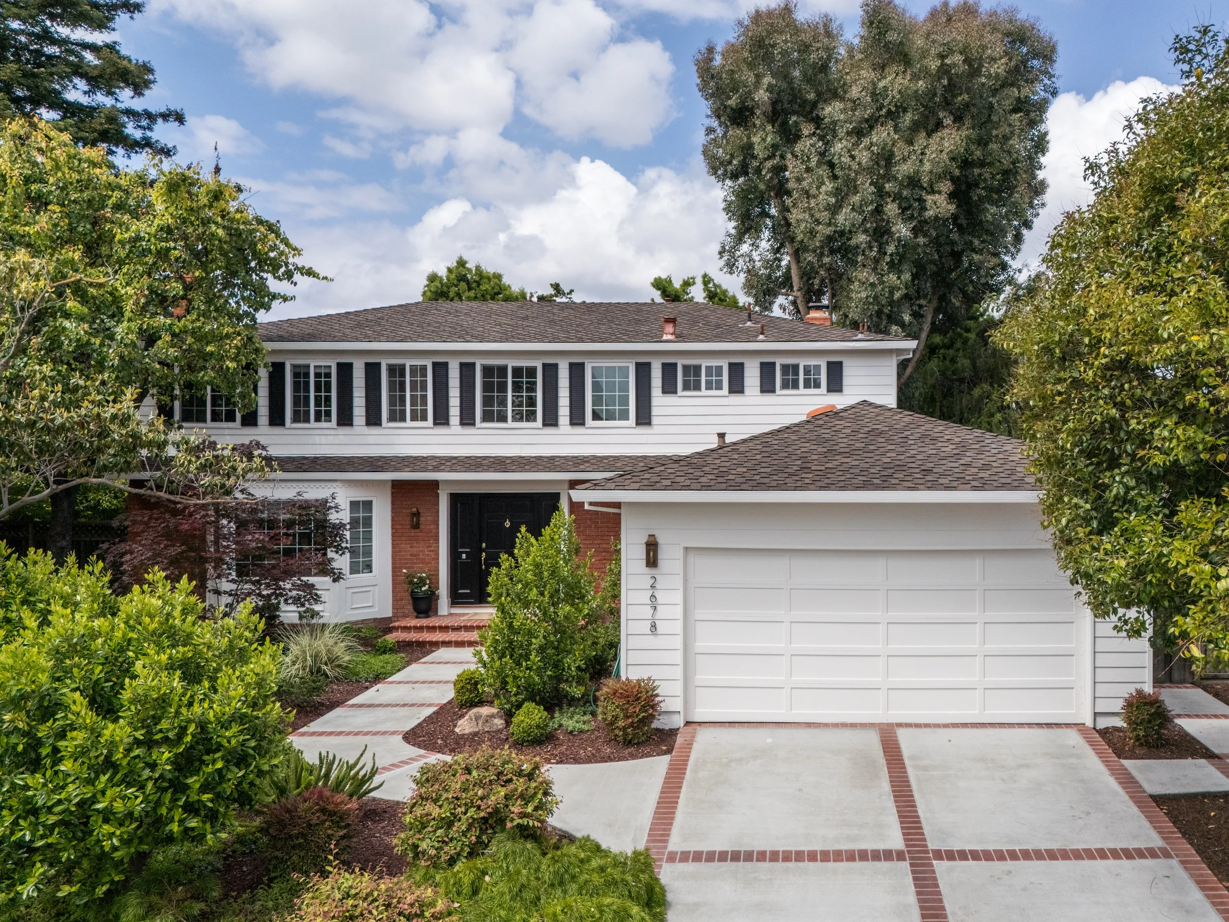 Front view of a white suburban house with a garage, black front door, and brick accents, surrounded by lush greenery and trees, under a partly cloudy sky.