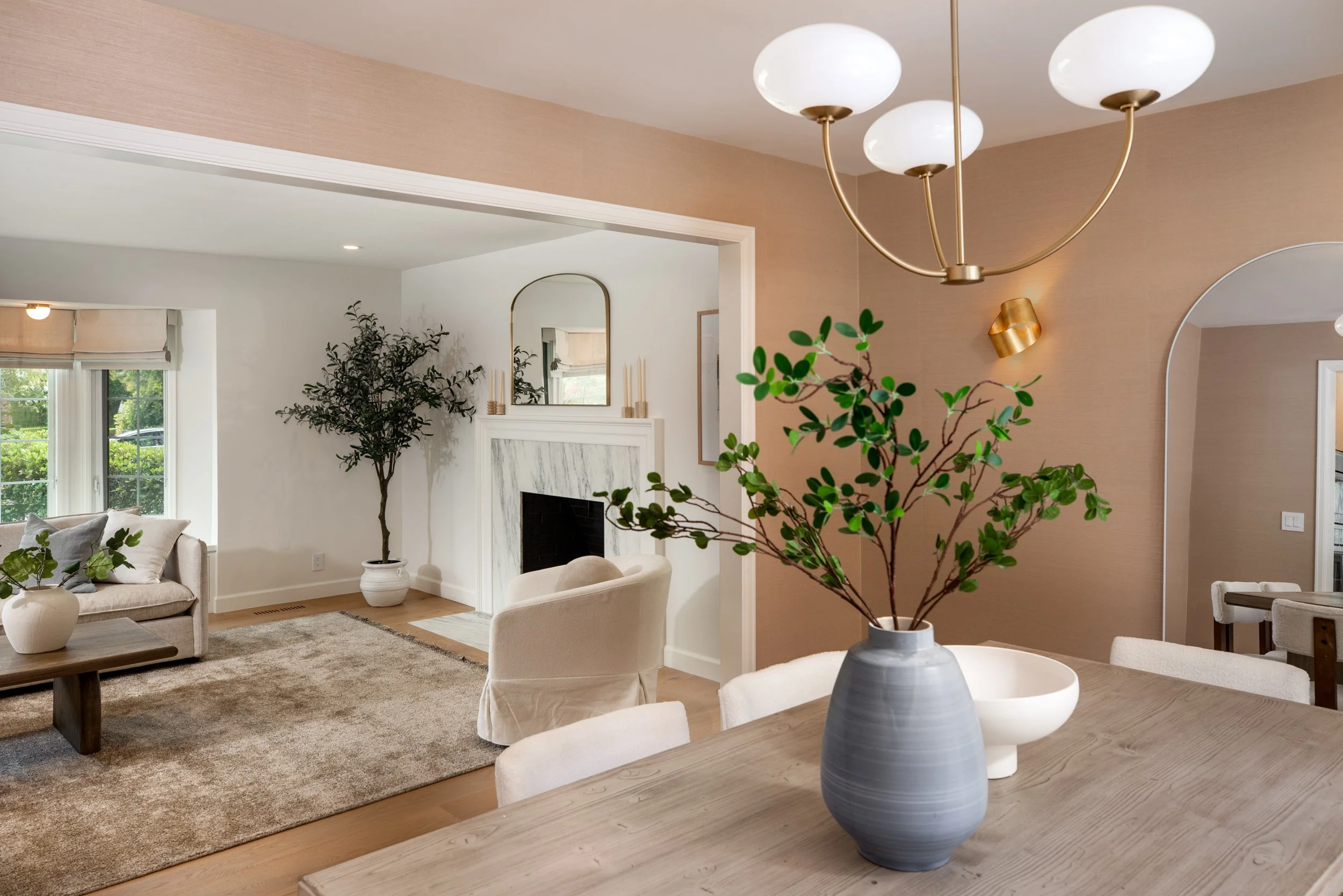 A living room and dining area with beige walls, wooden flooring, and modern decor. The living room has a beige sofa, a carpet, and a large window with blinds. The dining area features a wooden table with a large gray and white ceramic vase holding le