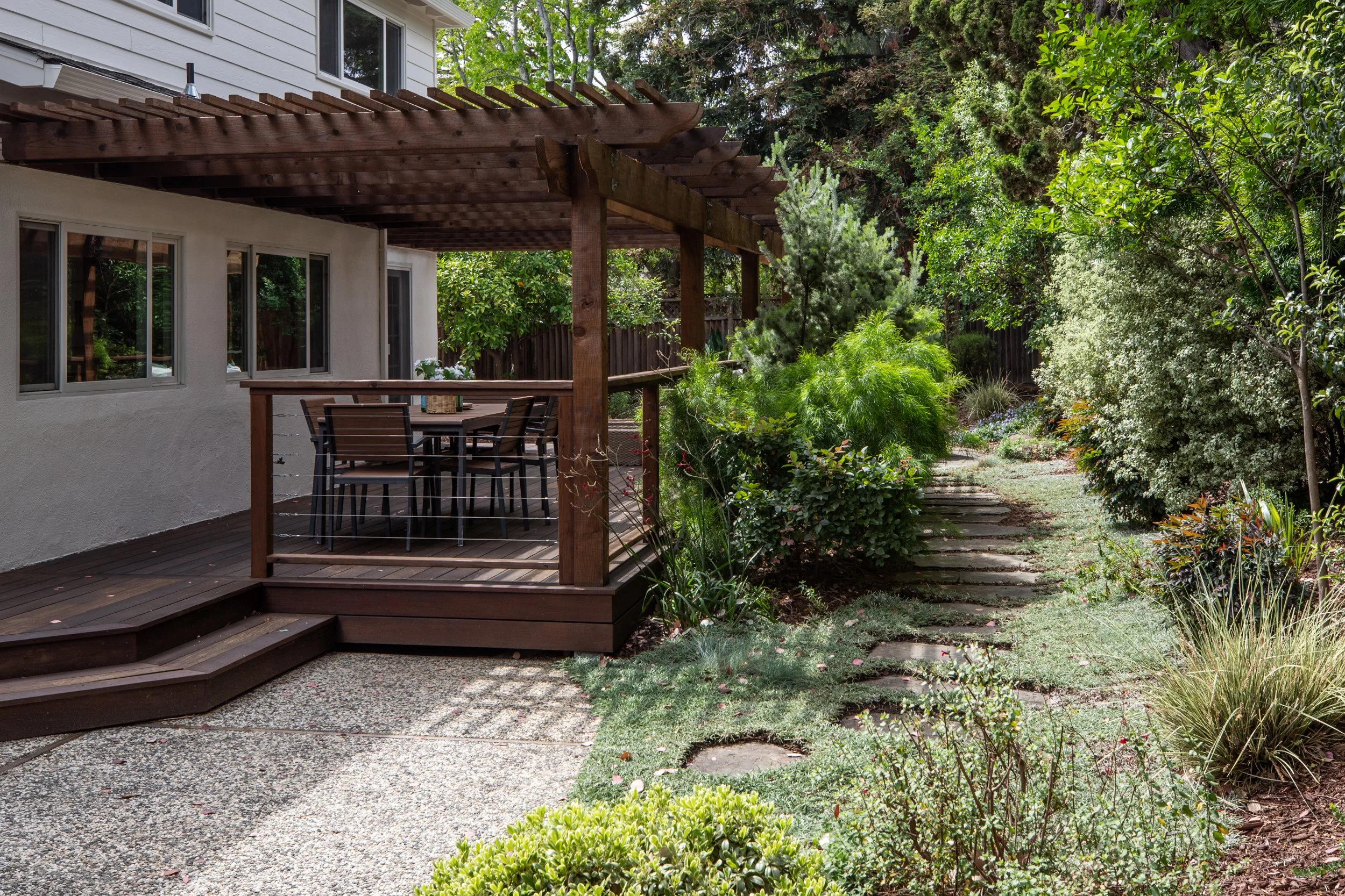 A backyard with a wooden deck and a pergola, surrounded by lush greenery and a stone pathway.