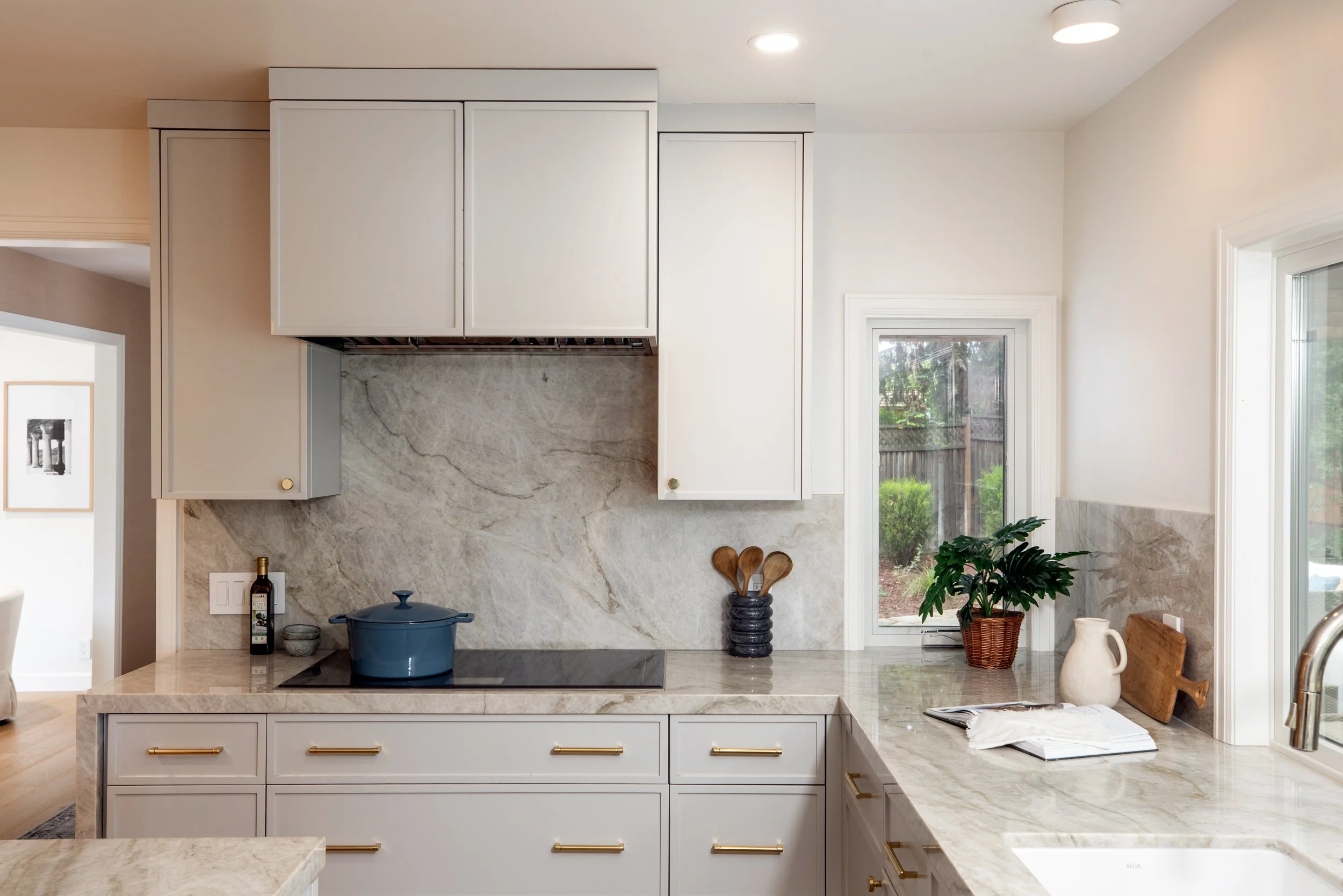 Modern kitchen with white cabinets, marble countertops, and large window overlooking a backyard with greenery.