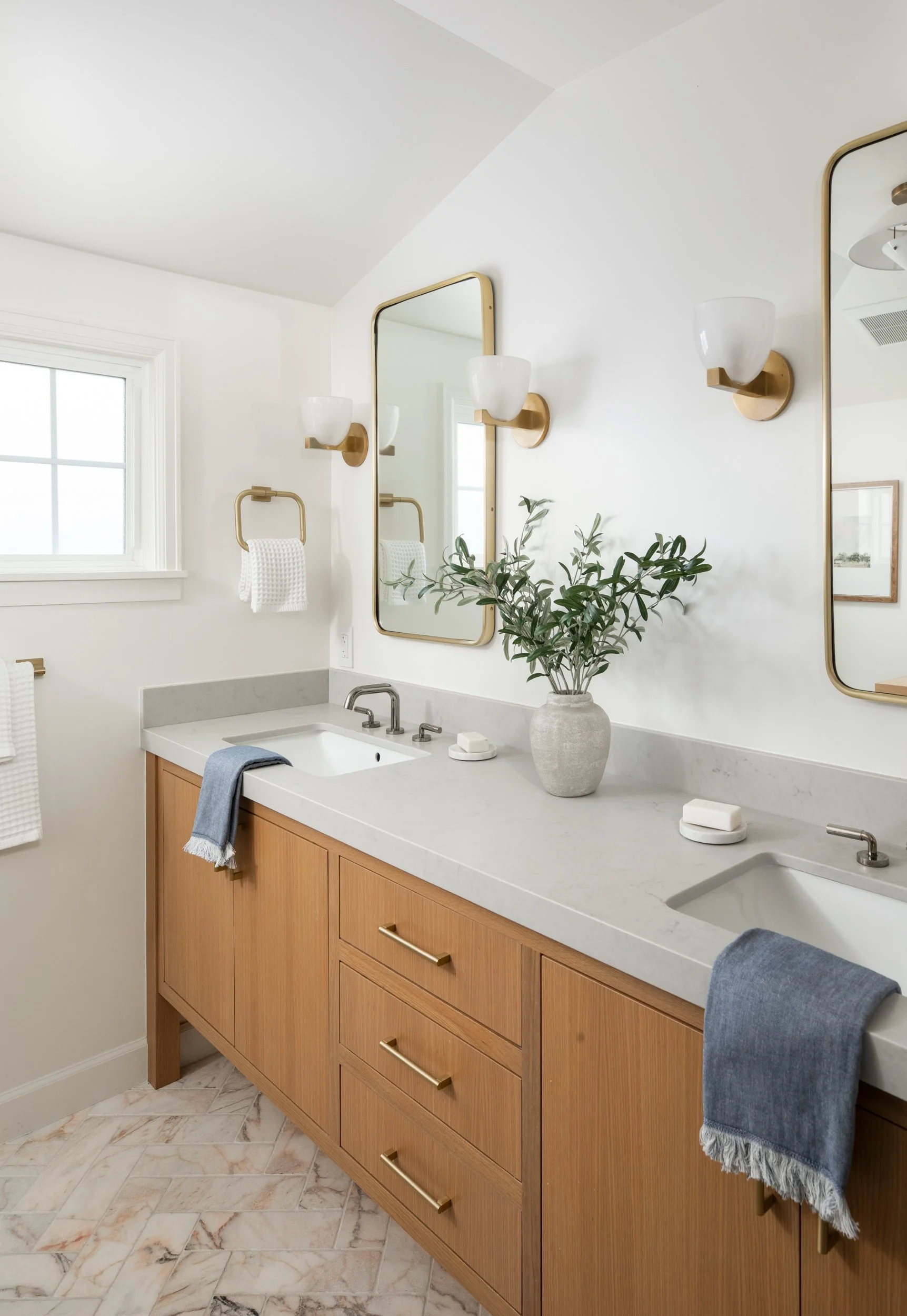 A bathroom vanity with a white countertop, wooden cabinets with gold handles, and a large mirror. There are two wall-mounted light fixtures, a window, a potted plant, soap, and blue towels hanging on the sides.