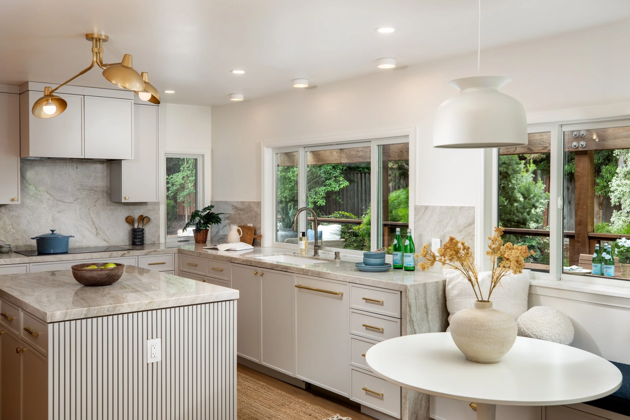 A bright, modern kitchen with white cabinets, marble countertops, and large windows overlooking a lush green yard. Pendant and recessed lighting enhance the space, which is decorated with a bowl of apples, plants, and bottles of sparkling water.