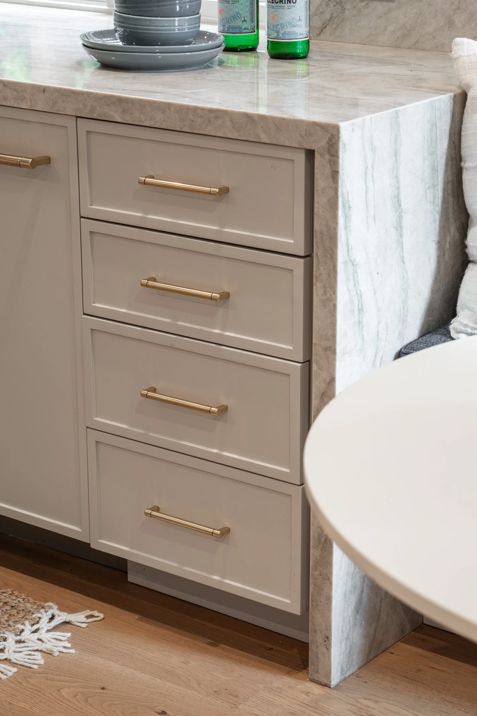 A kitchen countertop with stacked gray dishes and bowls, two green bottles of San Pellegrino sparkling water, and a beige marble-like side panel.