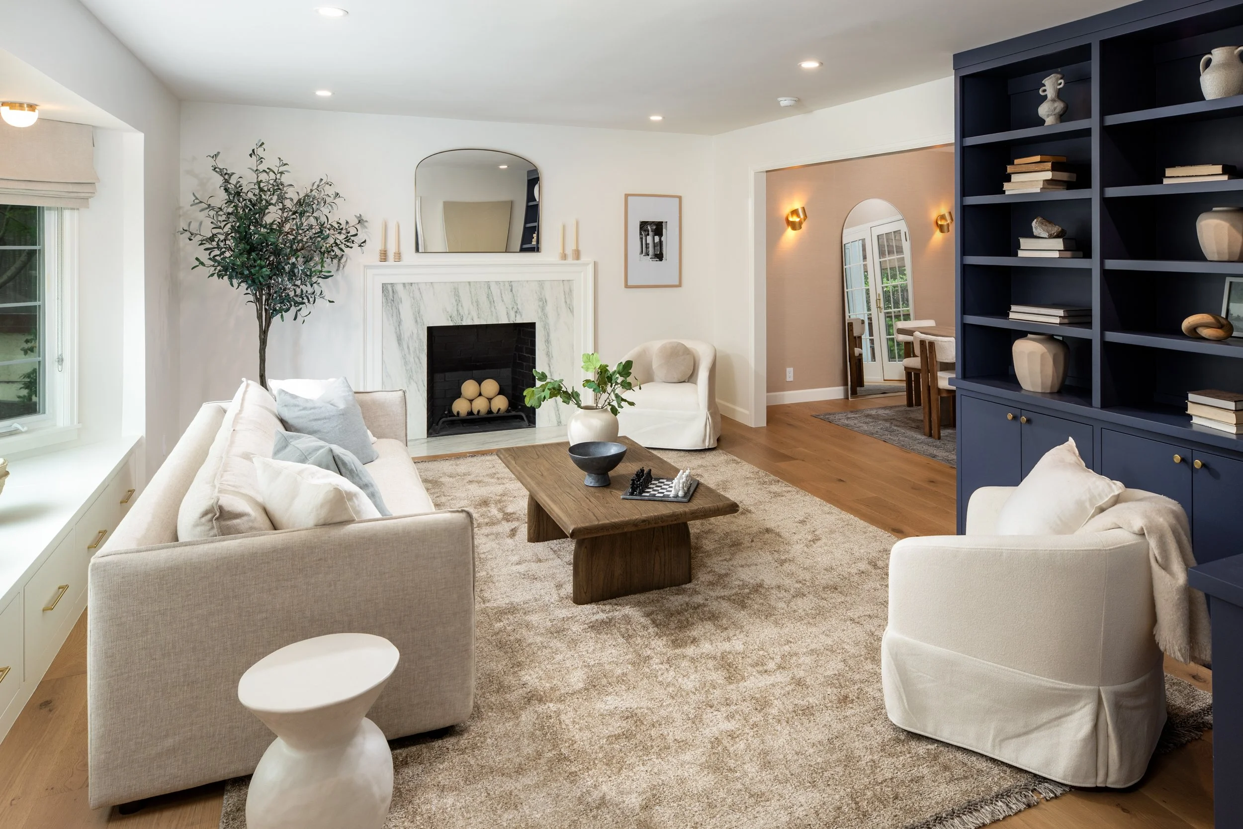 A living room with a beige sofa and armchair, a gray shaggy rug, a wooden coffee table, a dark blue bookcase with decorative items, a fireplace with a marble surround, a large window with white trim, a nearby dining area with a pink accent wall, and 