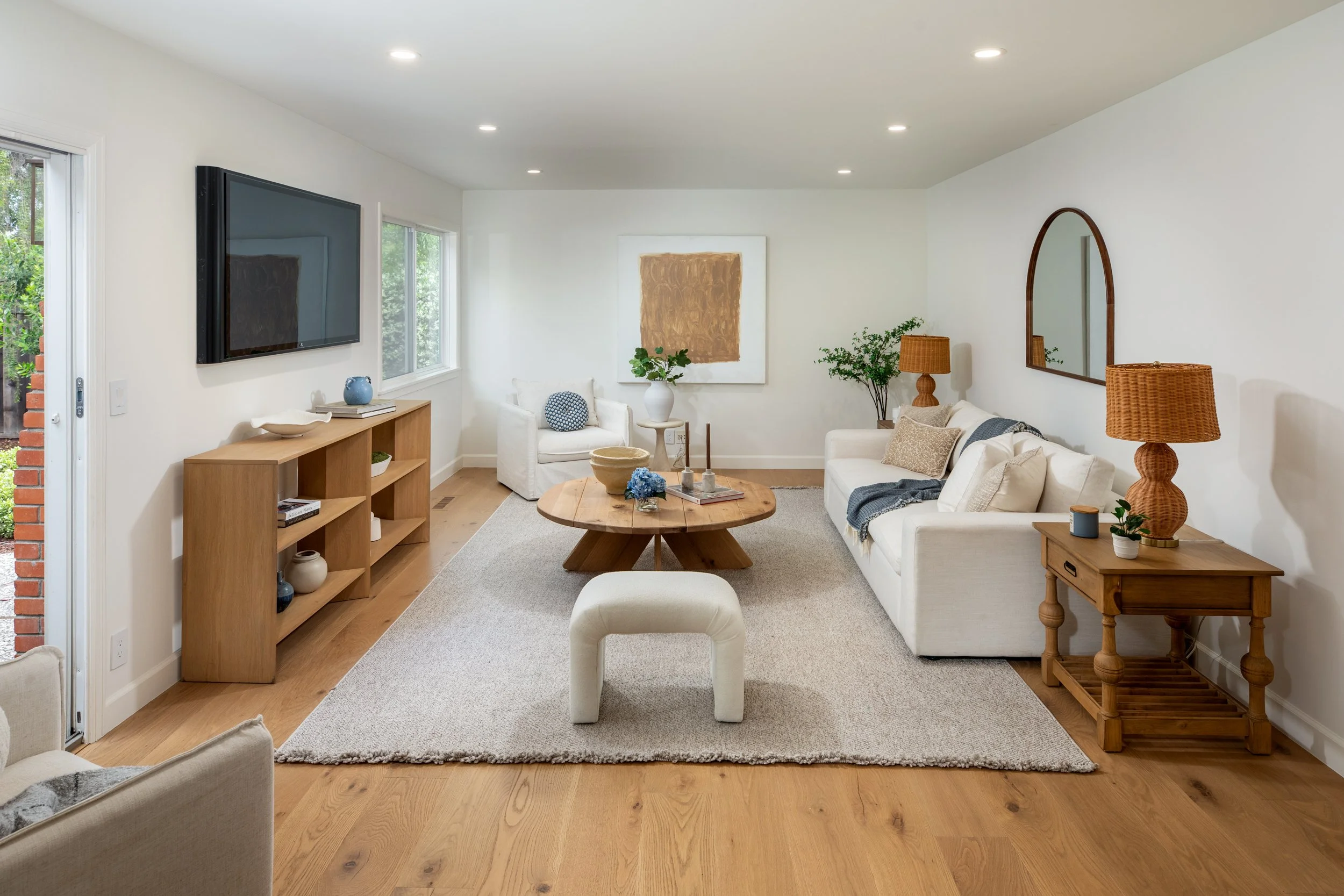 Living room with white walls, hardwood floor, beige rug, white sofas, wooden side tables, a wooden coffee table, wall-mounted TV, window with greenery outside, and decorative items like vases and lamps.