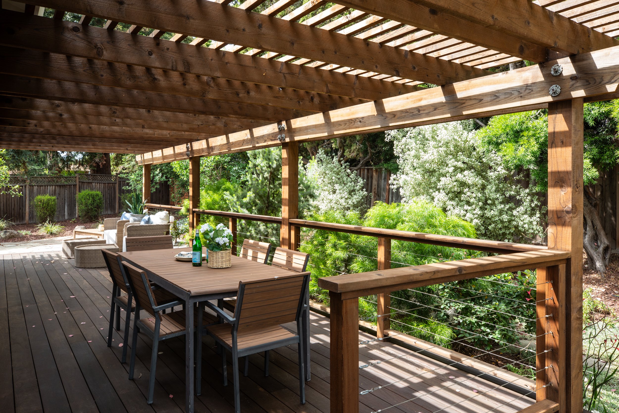 Wooden deck with an overhead pergola, patio furniture including a table and chairs, surrounded by lush green trees and plants.