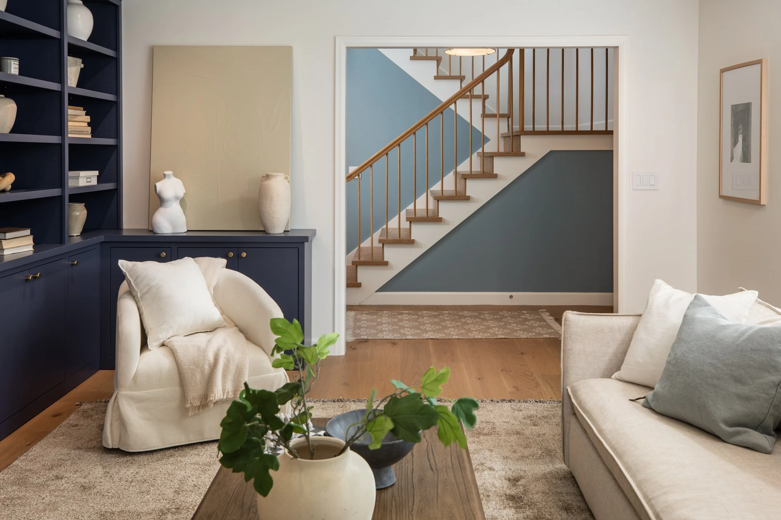 Living room with beige and gray furniture, a wooden staircase with a blue and gray wall behind it, and decorative vases on a blue cabinet.