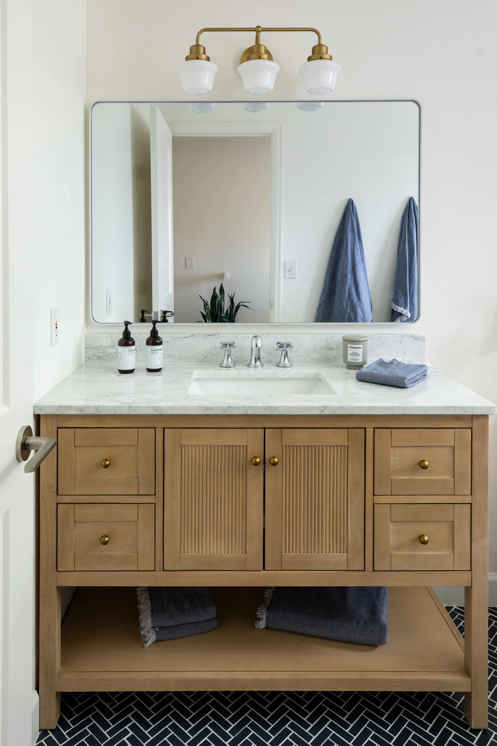 Bathroom vanity with a marble countertop, a large mirror, and a wooden cabinet. Two brown soap dispensers, a candle, and a folded towel are on the countertop. Two blue towels hang on the wall, and a plant is reflected in the mirror. The floor has bla