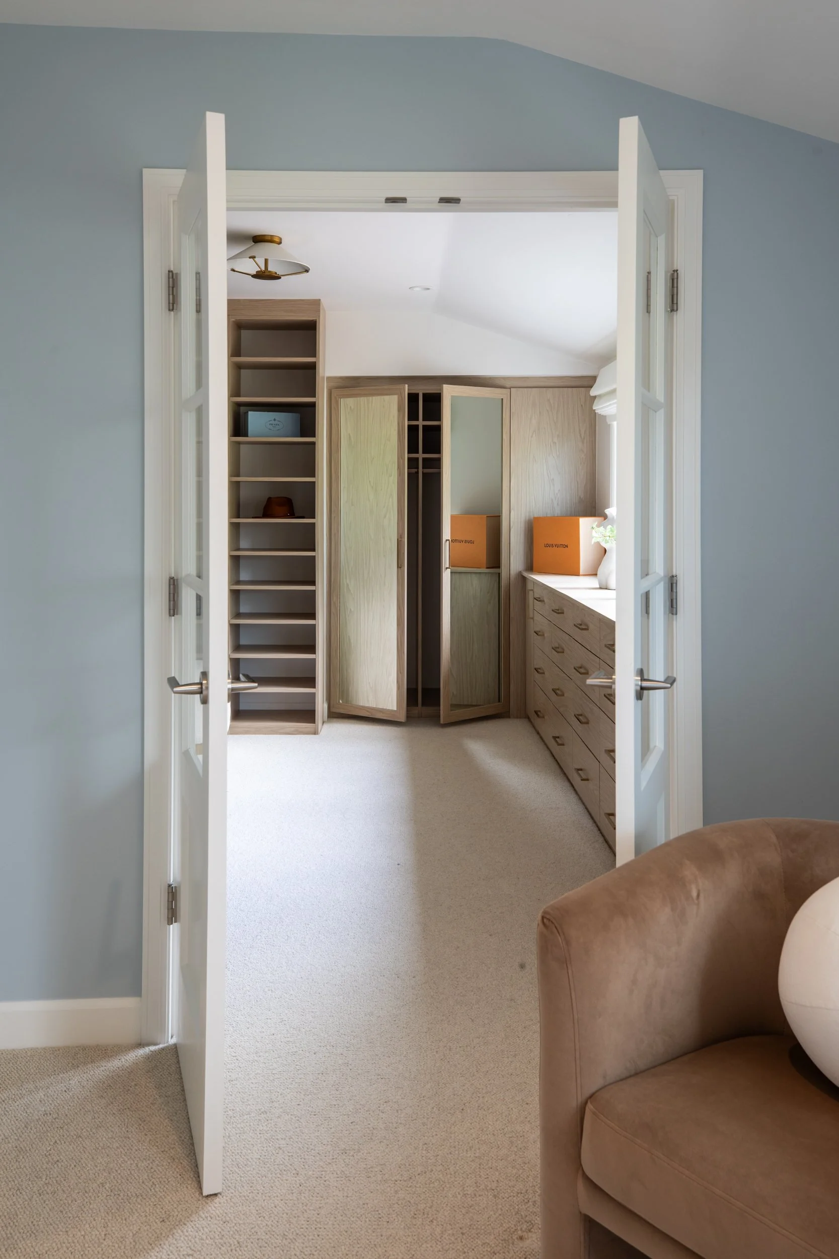 A doorway opens into a room with beige carpet, wooden shelves and cabinets, and a white ceiling. Two orange Louis Vuitton shopping bags are on a countertop next to a white decorative vase with flowers. In the foreground, part of a brown armchair is v