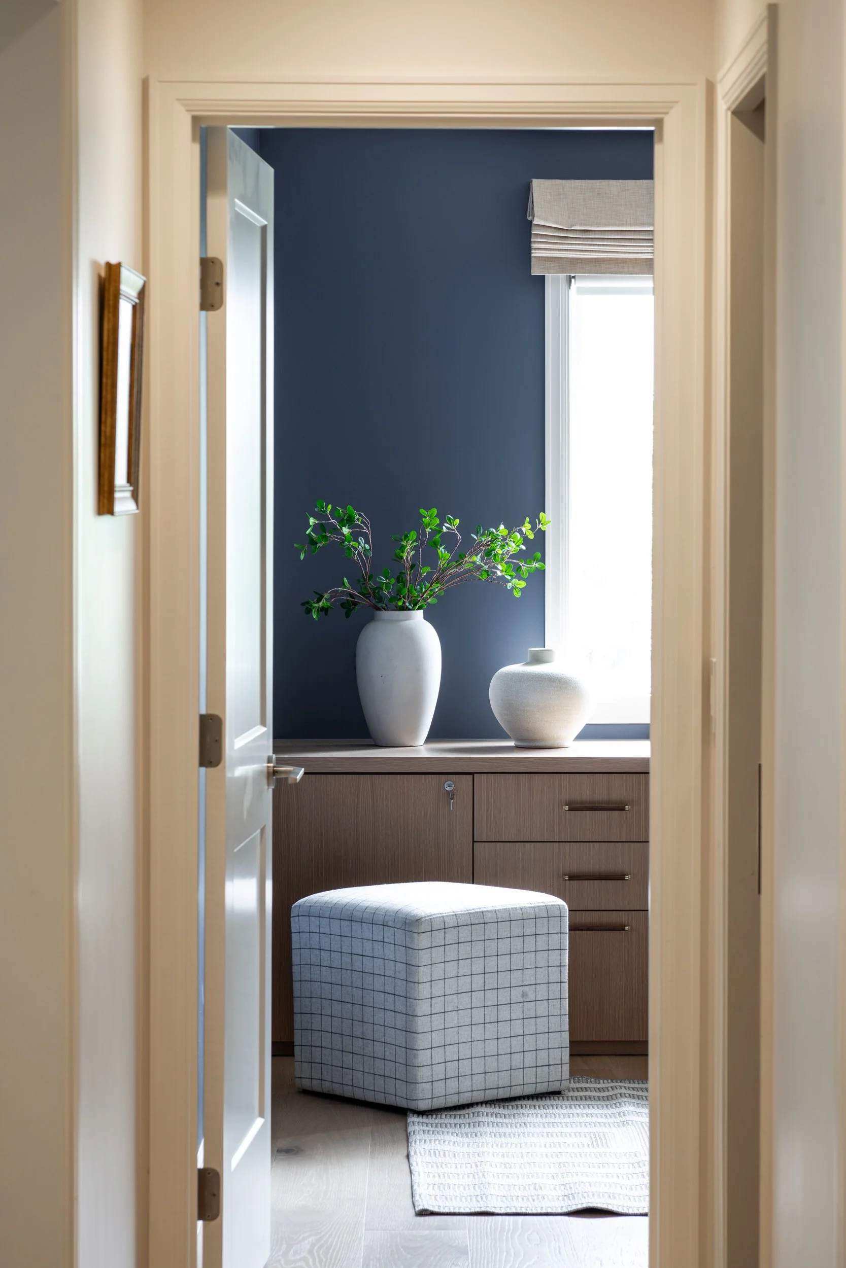 A view through a doorway into a room with a dark blue accent wall, a window with a beige Roman shade, a large white vase with green leaves, a smaller white ceramic vase, a wooden cabinet, and a plaid ottoman on a rug.