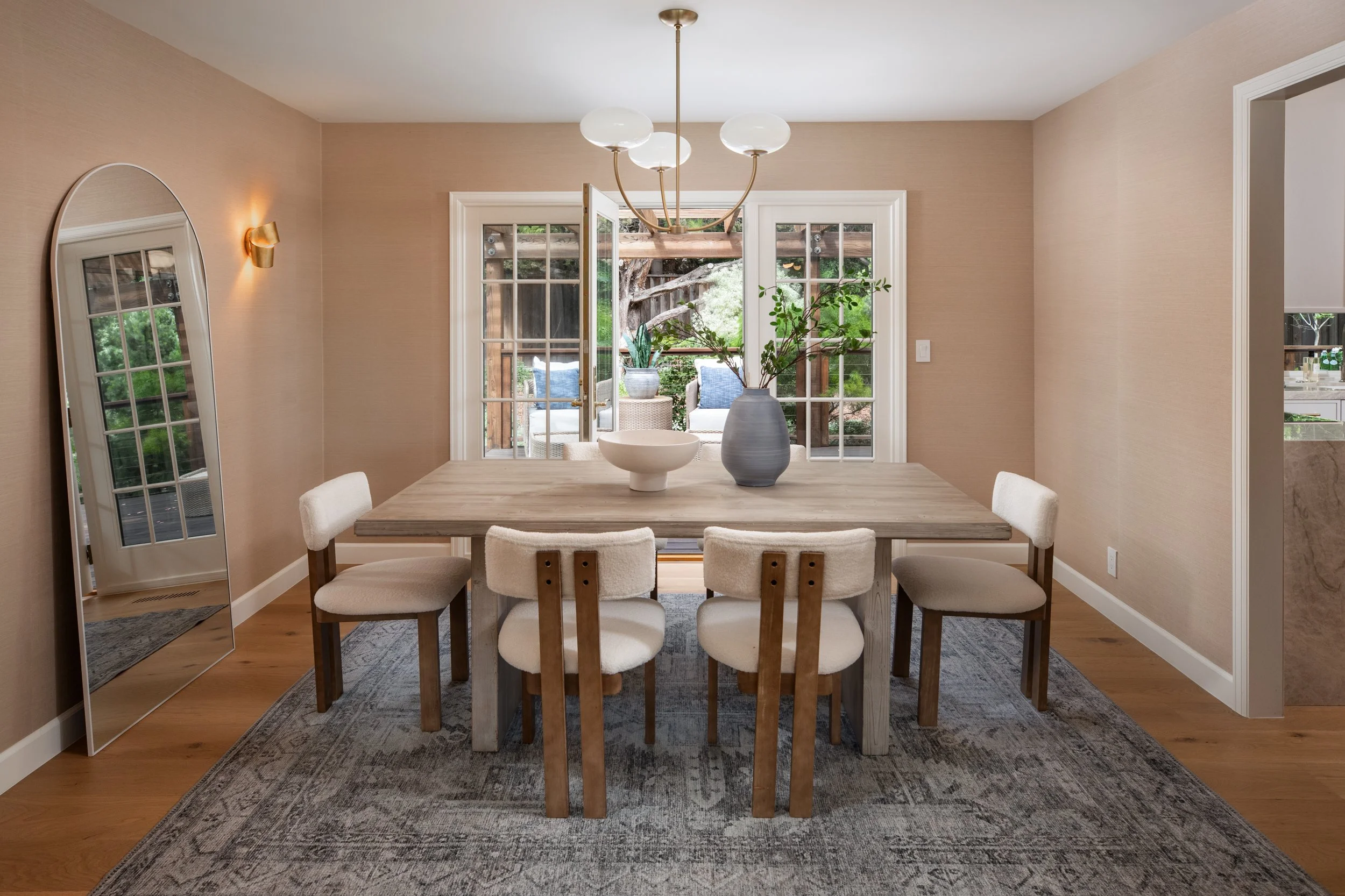 Dining room with wooden table, six white upholstered chairs, a large vase with green branches, a modern chandelier, a large mirror, and glass doors leading to an outdoor patio with seating and trees.