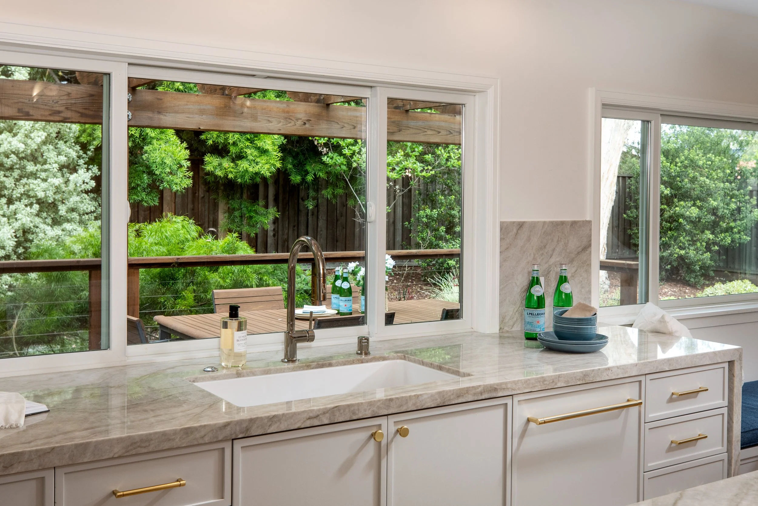 Kitchen view through a large window, showing a wooden deck with greenery outside, and a countertop with bottles of San Pelligrino water, bowls, and a hand soap dispenser inside.