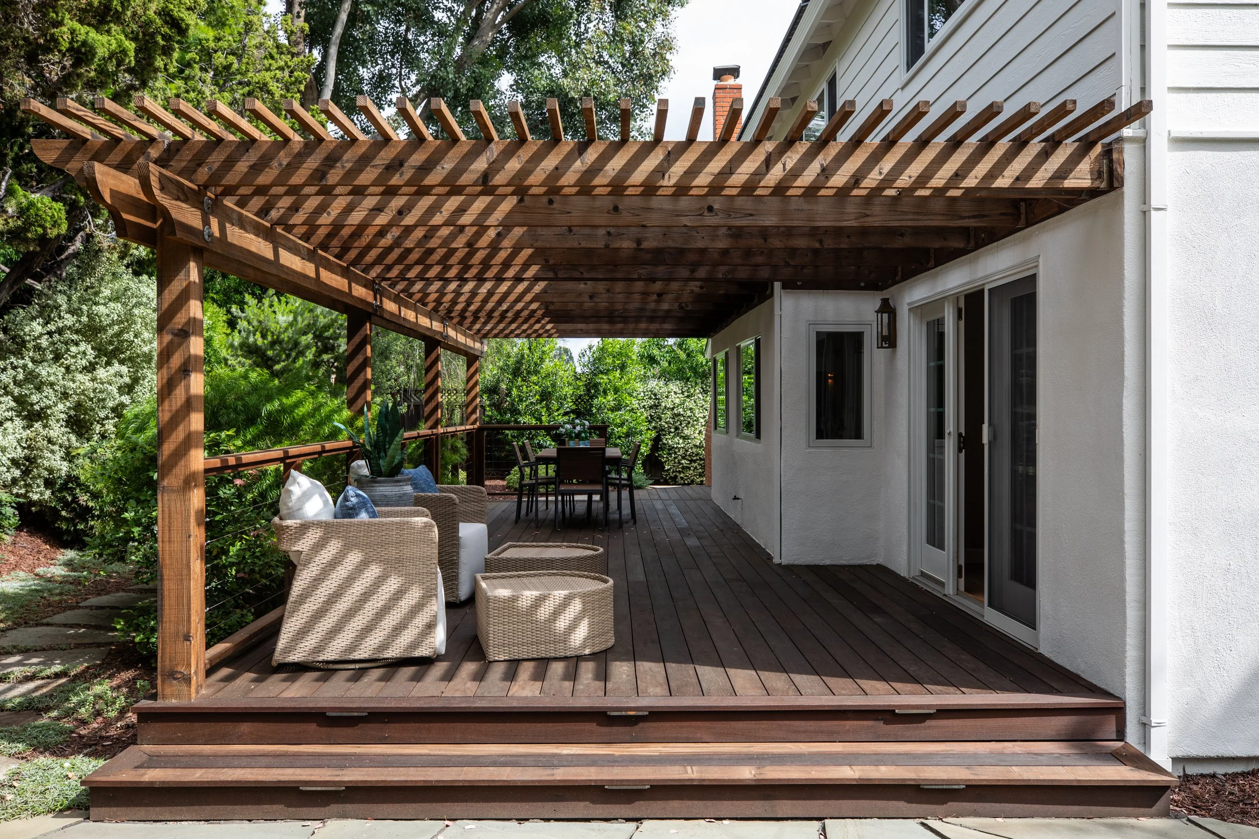 Wooden deck extension with outdoor furniture, including wicker chairs and a dining table, attached to a white house.