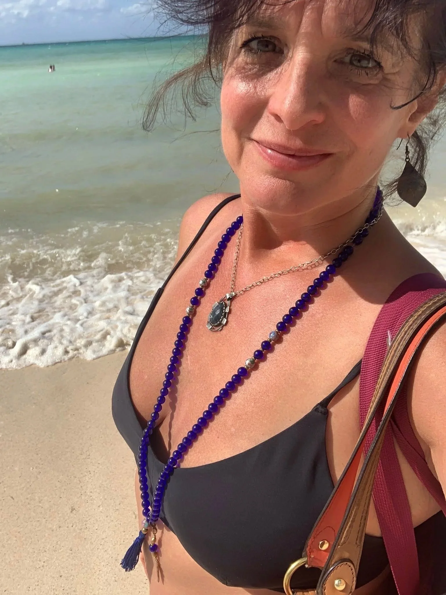 A woman with short curly hair and gray eyes smiling, standing on a beach with the ocean and sky in the background, wearing a black top and layered necklaces, with a maroon strap bag over her shoulder.