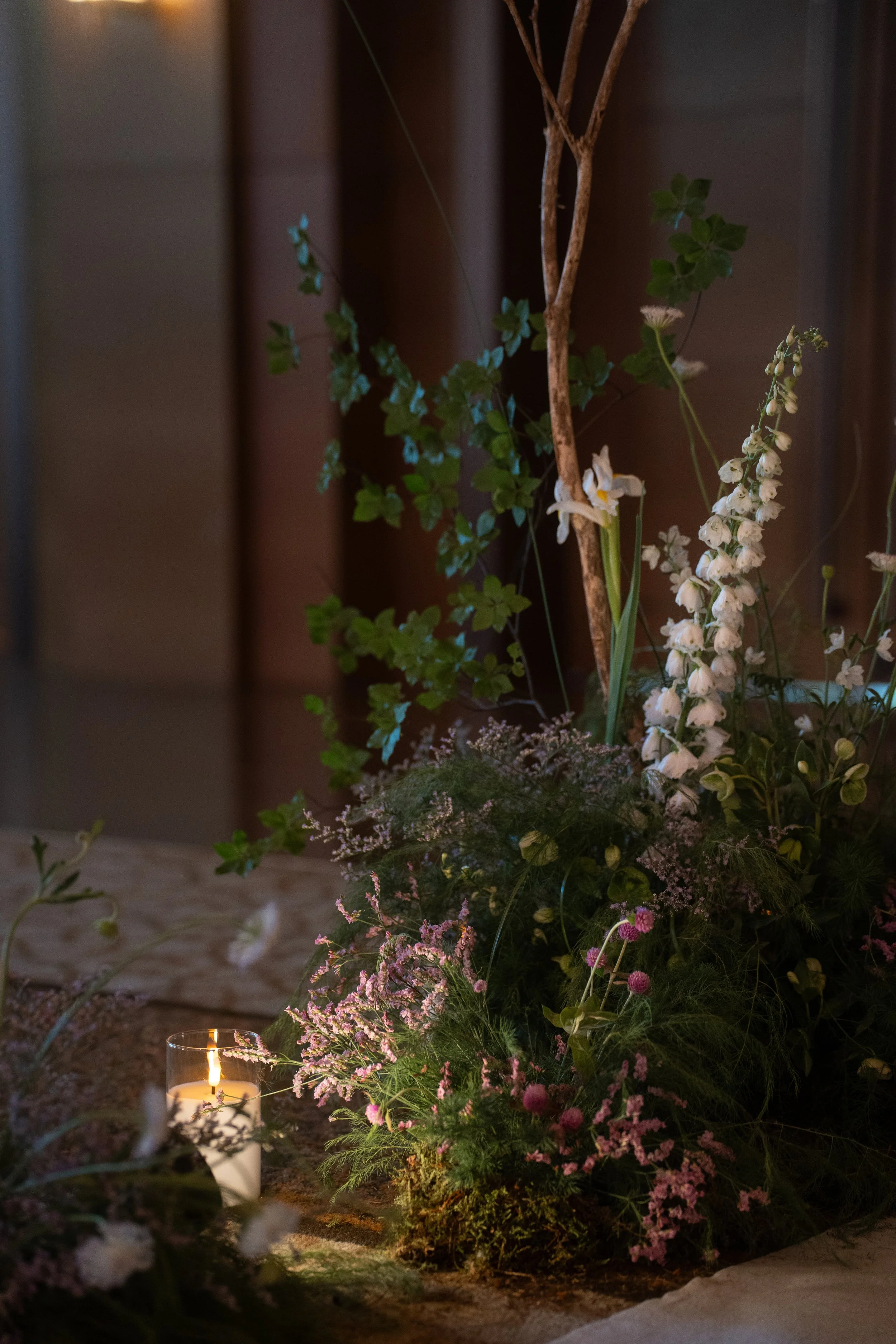 A floral arrangement with pink and white flowers, green foliage, and a candle in a glass holder is situated on the ground next to a brown wooden wall.