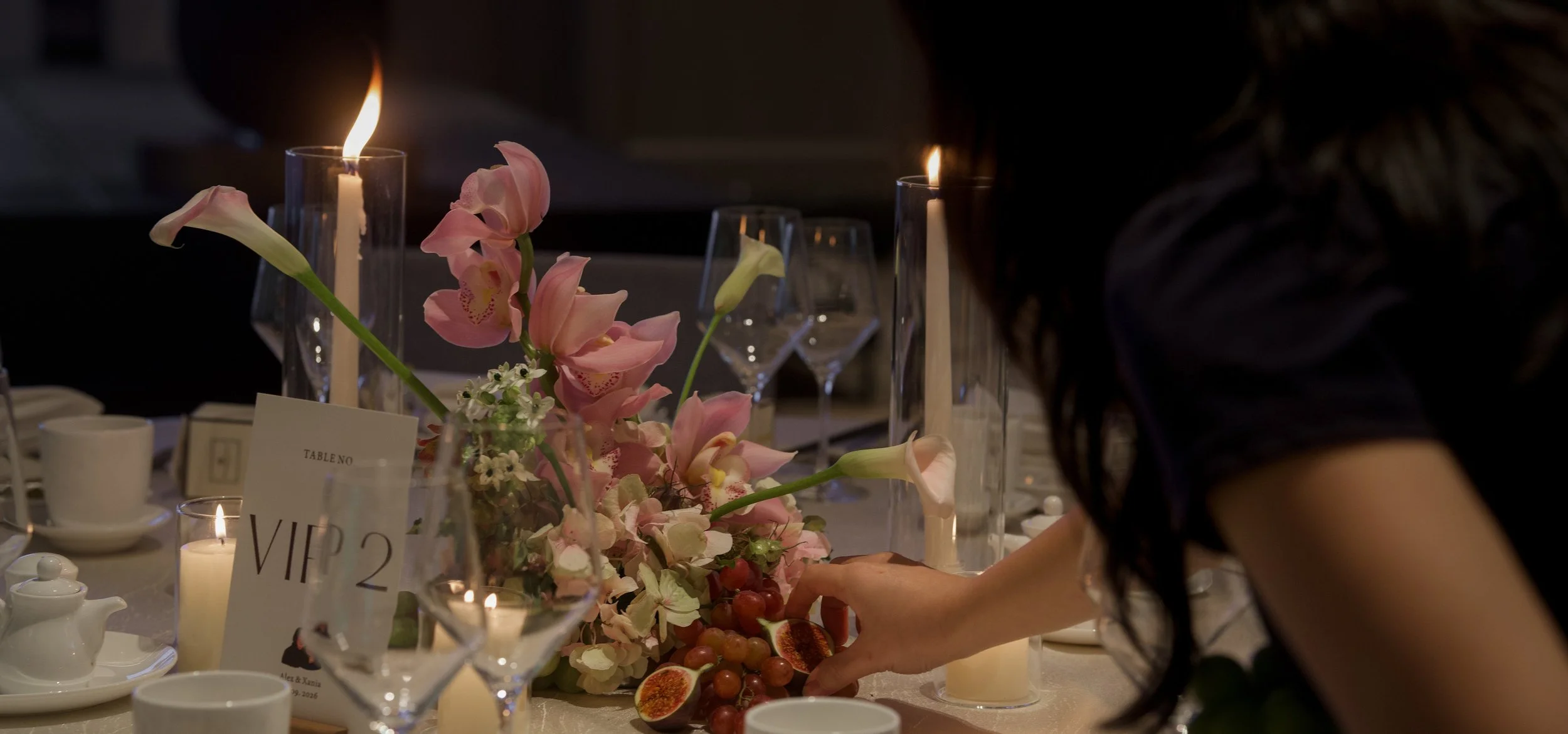 Florist arranging a candlelit wedding tablescape with pink cymbidium orchid flowers and fresh fruit like grapes and figs.
