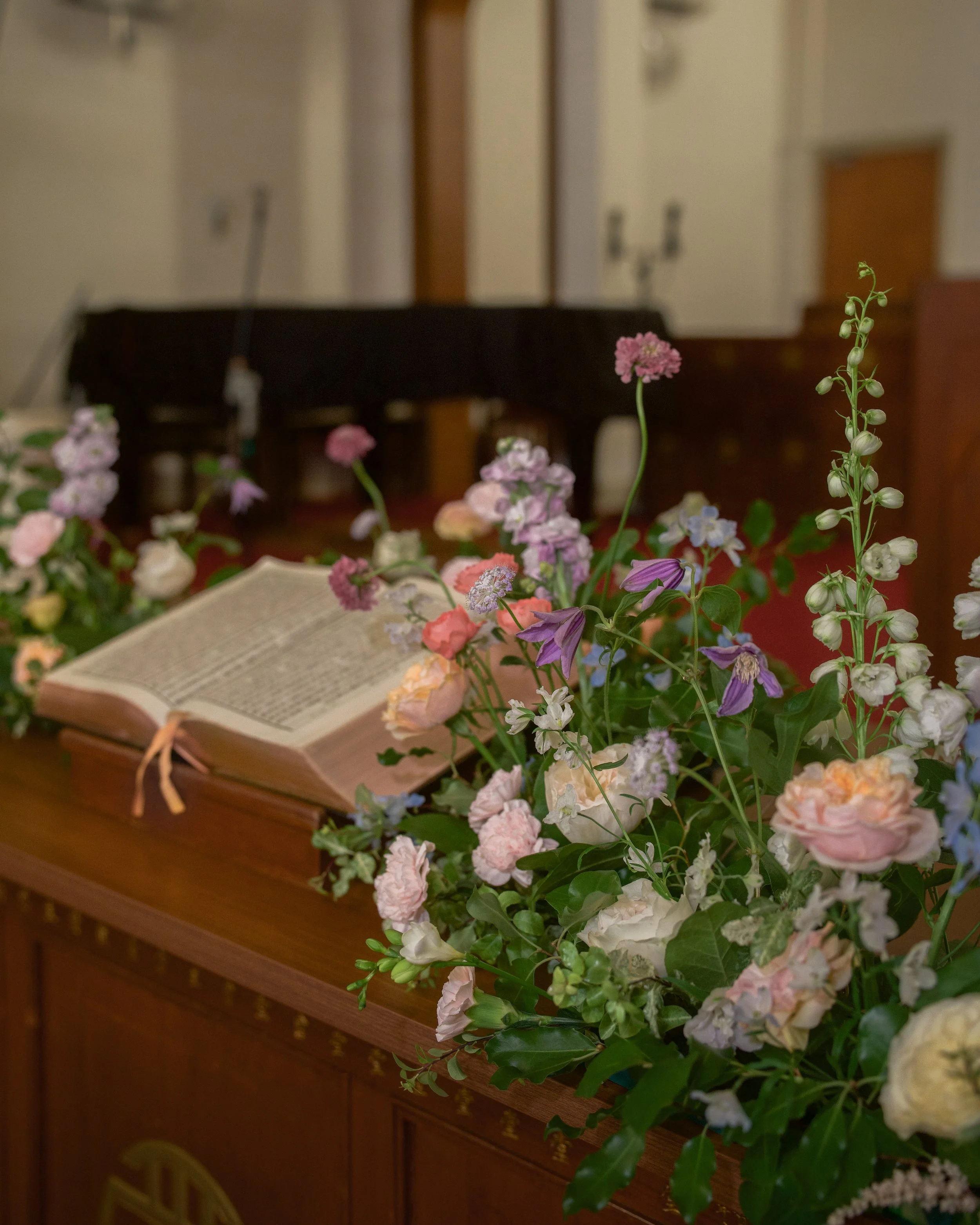 A bouquet of pink, purple, and white flowers with green leaves placed on a wooden surface, with an open book in the foreground. In the background, there is a dark-colored piano and a beige-colored wall with outlets.