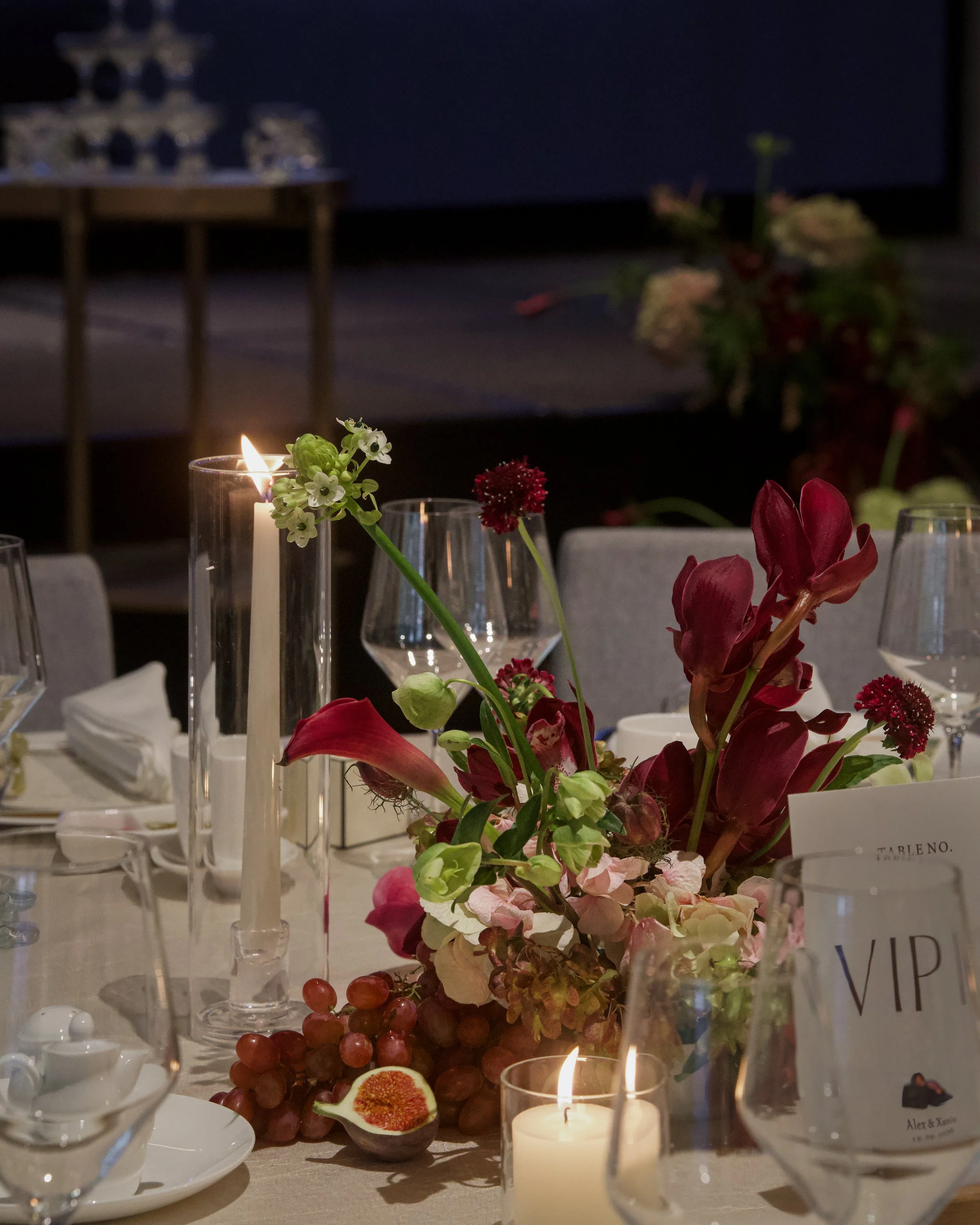 A close-up of a table with floral arrangements, candles, and wine glasses, set for an elegant dinner event. The background shows a blurry dark setting with more floral decorations.