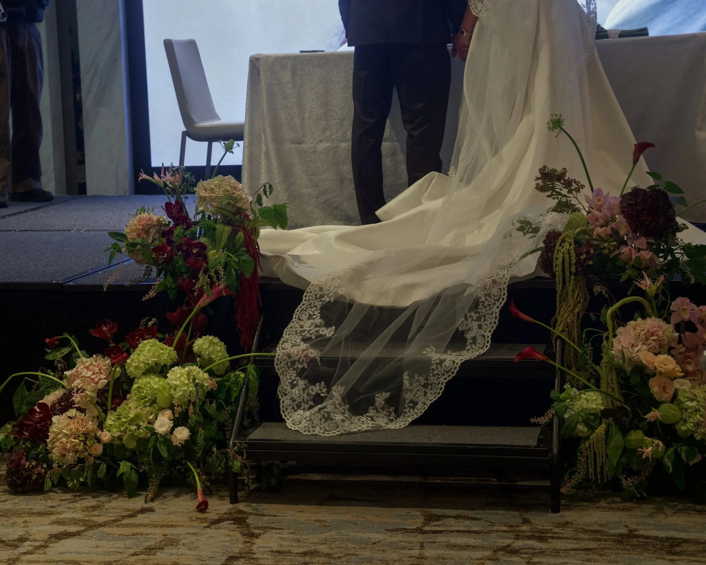 A couple holding hands during their wedding ceremony, standing behind a table and surrounded by a variety of colorful flowers and greenery.