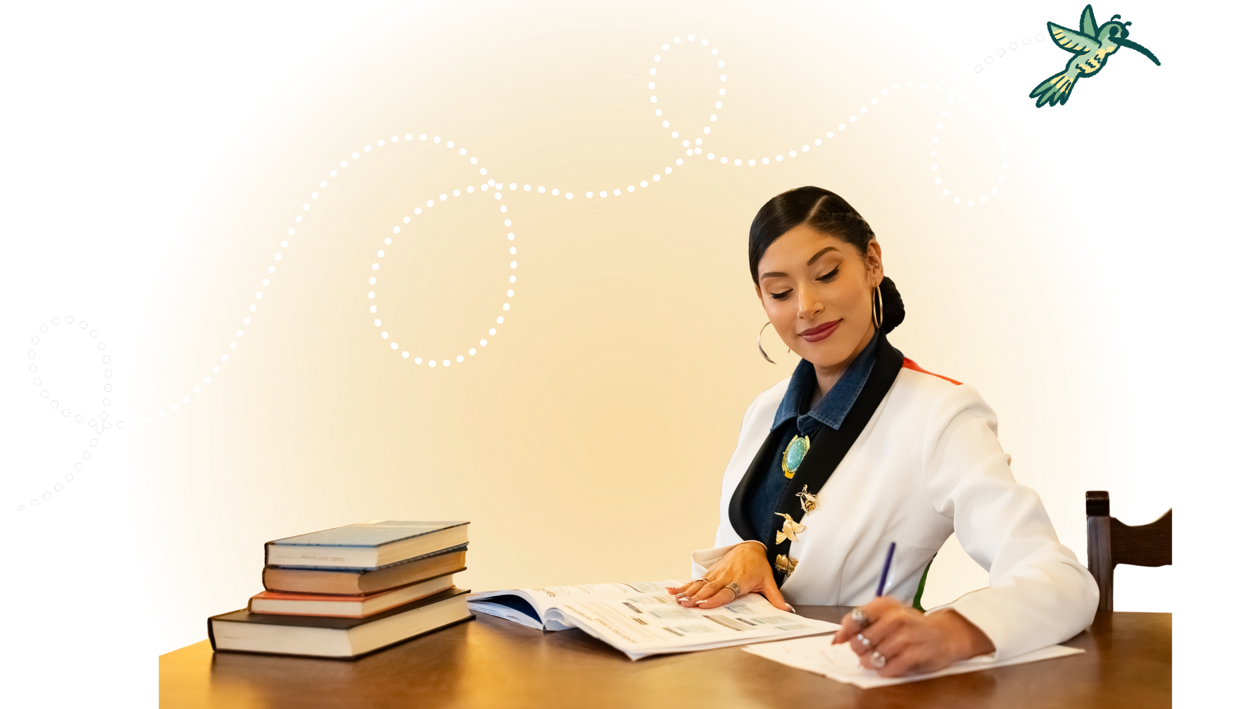 The author dressed in a white blazer with a large turquoise brooch, sitting at a wooden table, reading a magazine, with several stacked books and notebooks nearby. She has dark hair styled in a side part and is wearing large hoop earrings.