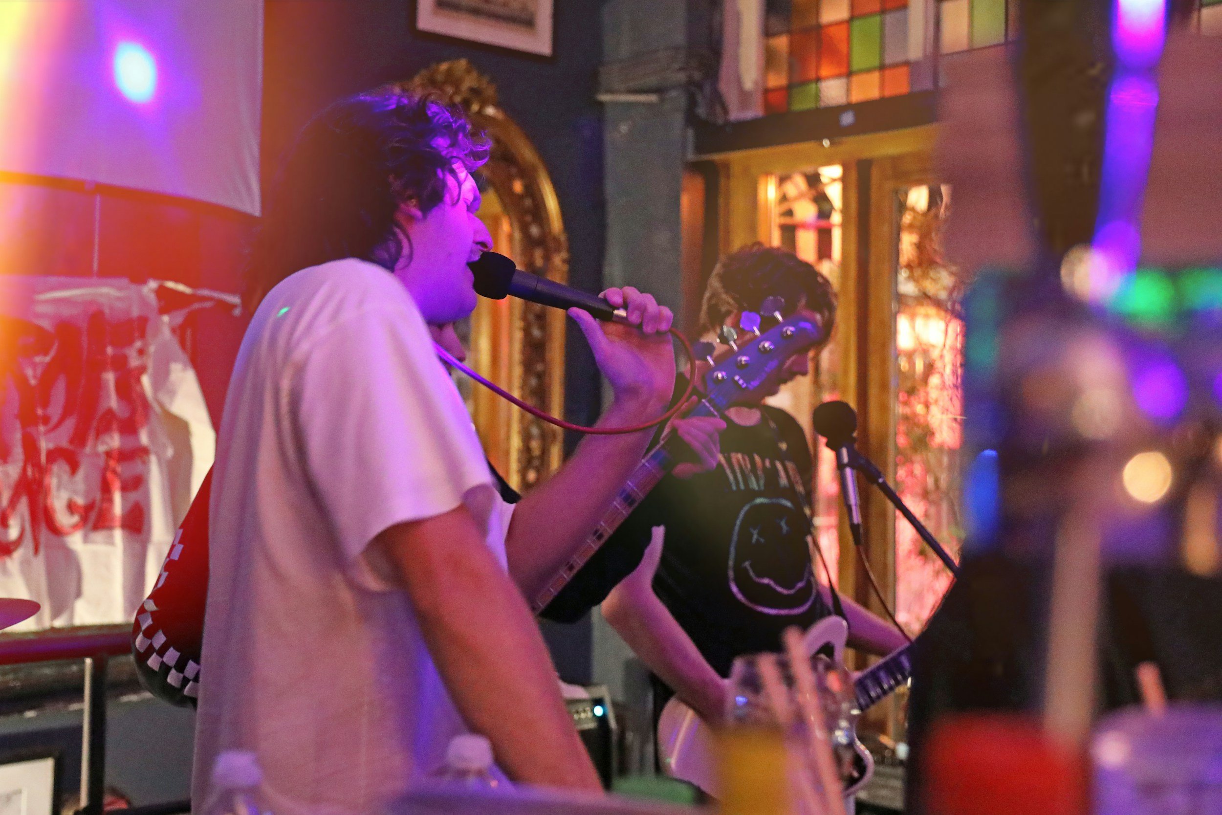 Two people playing guitars in a dimly lit room with colorful lights, one person with curly hair singing into a microphone.