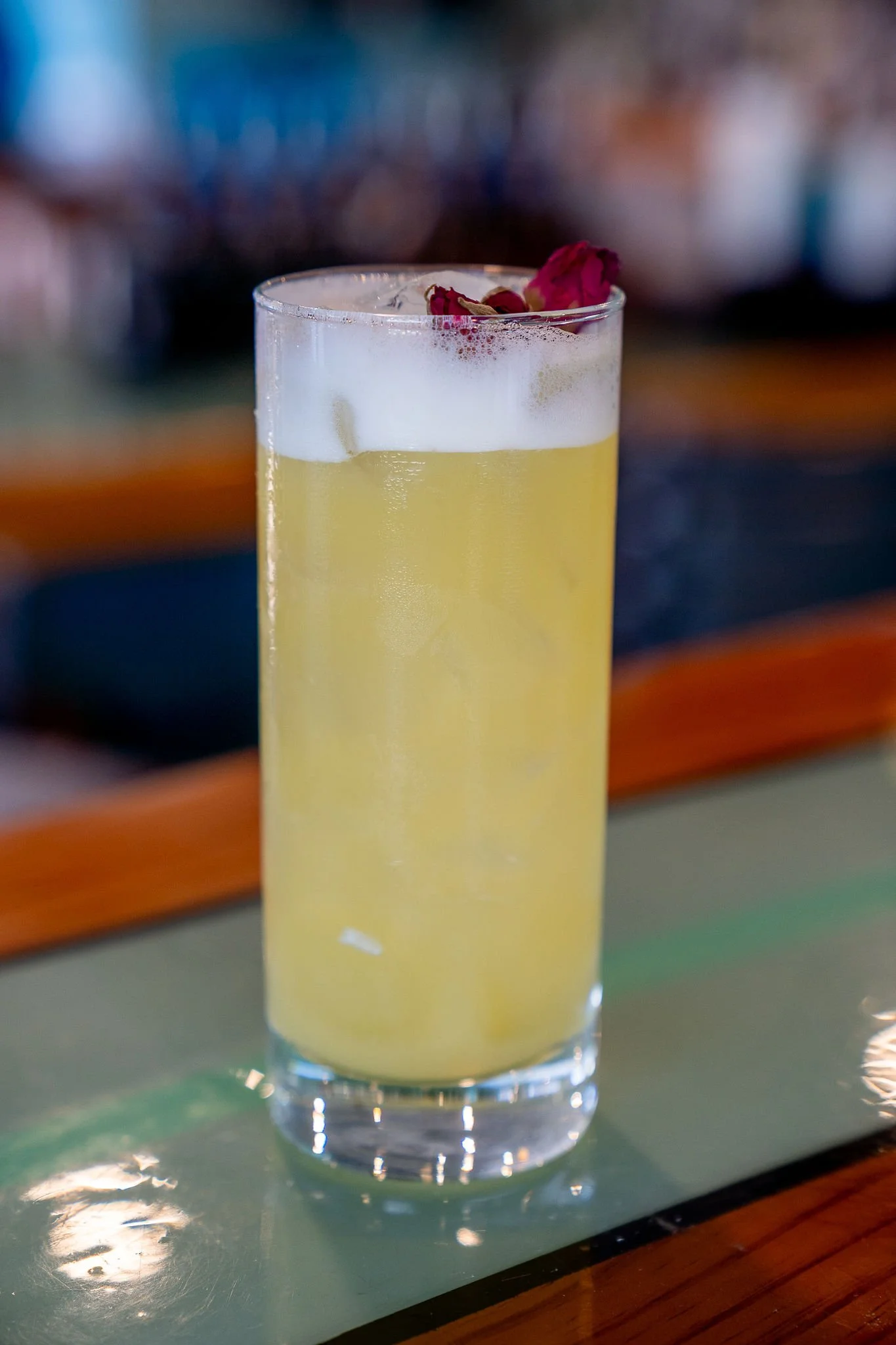A tall glass of yellow beverage with foam and dried roses on top, placed on a reflective surface.