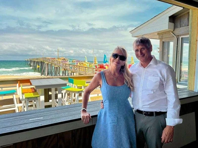 A woman in a blue dress and sunglasses and a man in a white shirt and gray pants are smiling and standing together indoors near a window, with a beach and pier visible in the background.