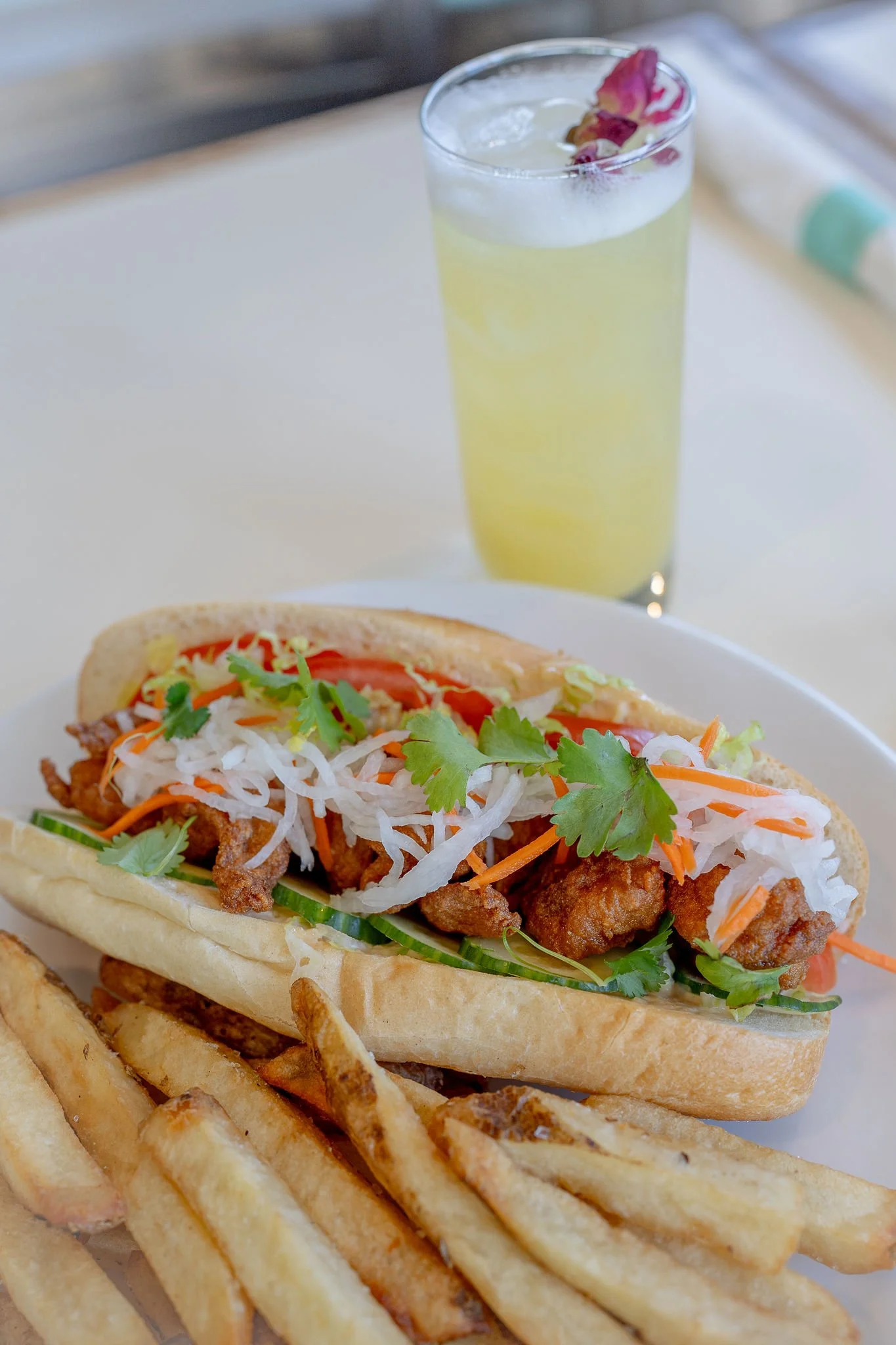 A hot dog in a bun topped with fried chicken, vegetables, fresh herbs, and shredded vegetables served with a side of waffle fries, with a glass of light-colored soda garnished with a flower petal in the background.
