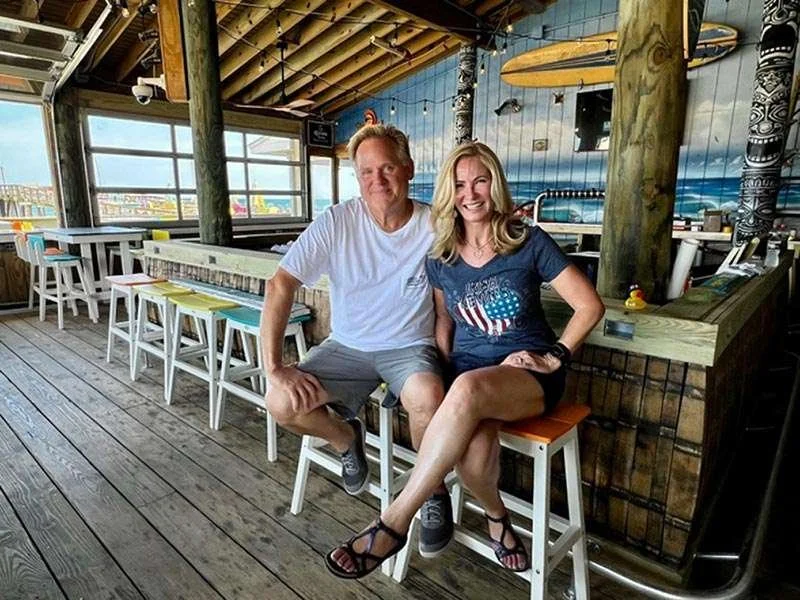 A man and woman sitting at a bar with surfboards mounted on the ceiling in the background, in a wooden decor setting, smiling at the camera.