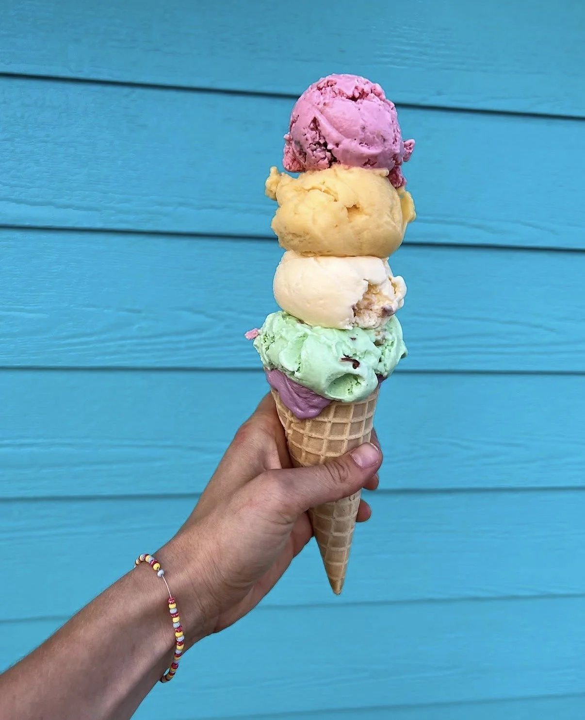 Hand holding a cone with four scoops of colorful ice cream against a blue wooden background.