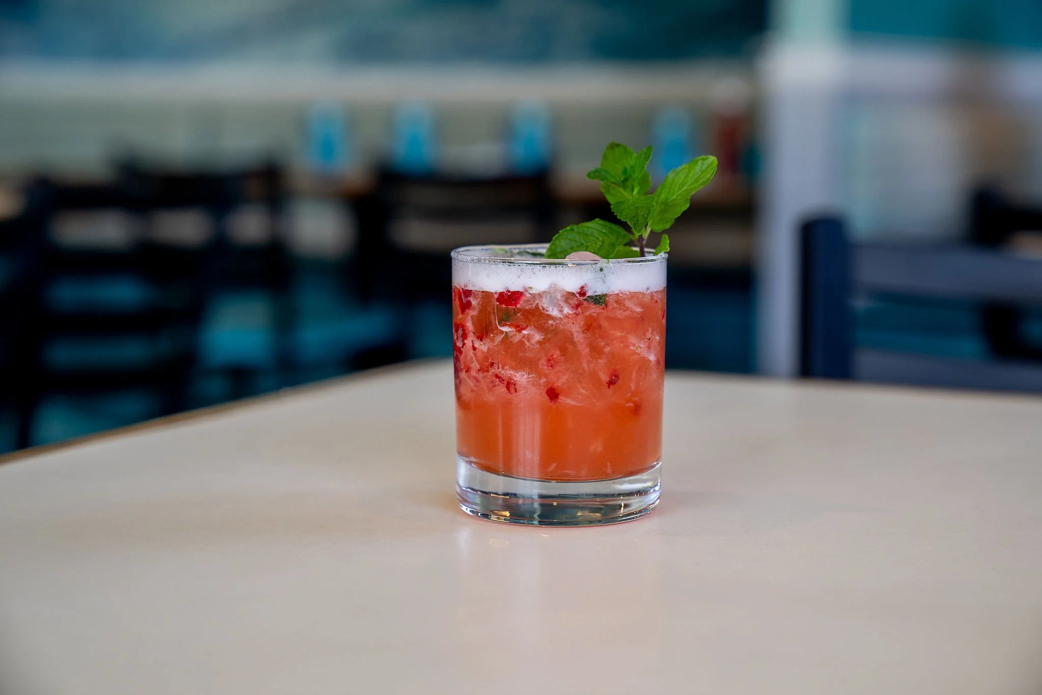 A glass of pinkish-red cocktail garnished with fresh mint leaves, placed on a white surface.