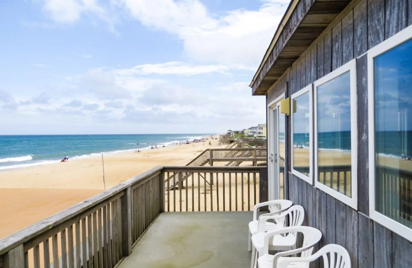 Balcony of a beach house with white plastic chairs overlooking a sandy beach and ocean under a partly cloudy sky.