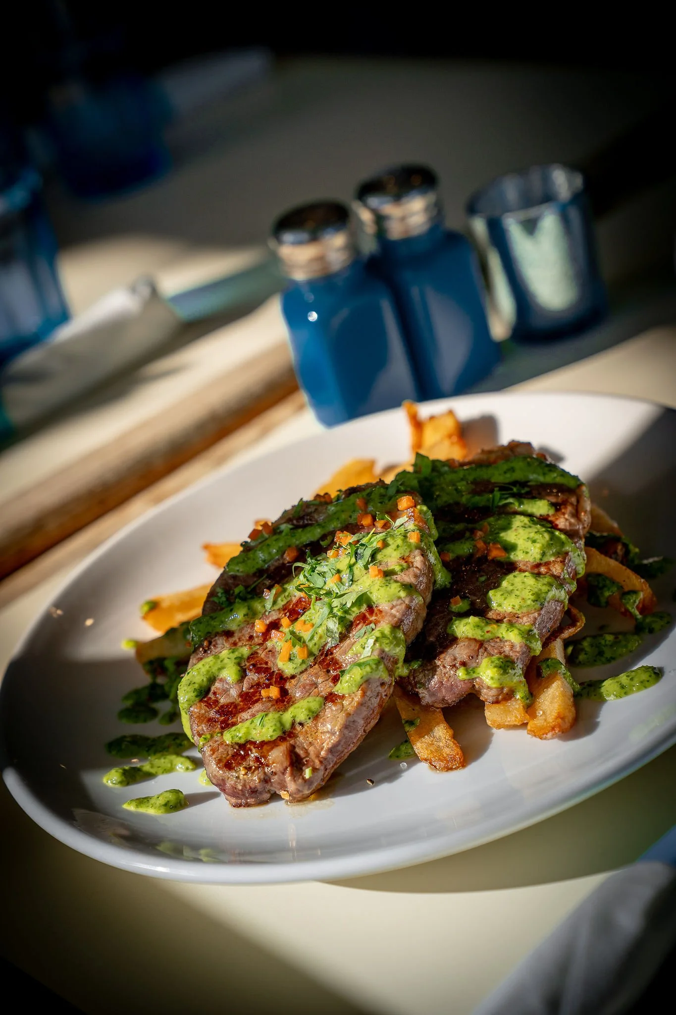 Close-up of a plate of steak topped with a green sauce, garnished with herbs and small diced vegetables, accompanied by crispy fries, with a blurred background of salt and pepper shakers.