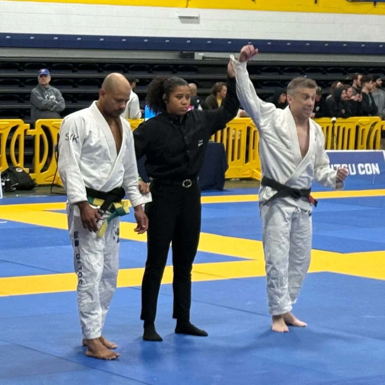 A female referee is raising the arm of a male Brazilian Jiu-Jitsu competitor wearing a white gi, black belt, and barefoot, who is celebrating with a clenched fist on a blue and yellow mat at a competition, with spectators and other competitors in the background.