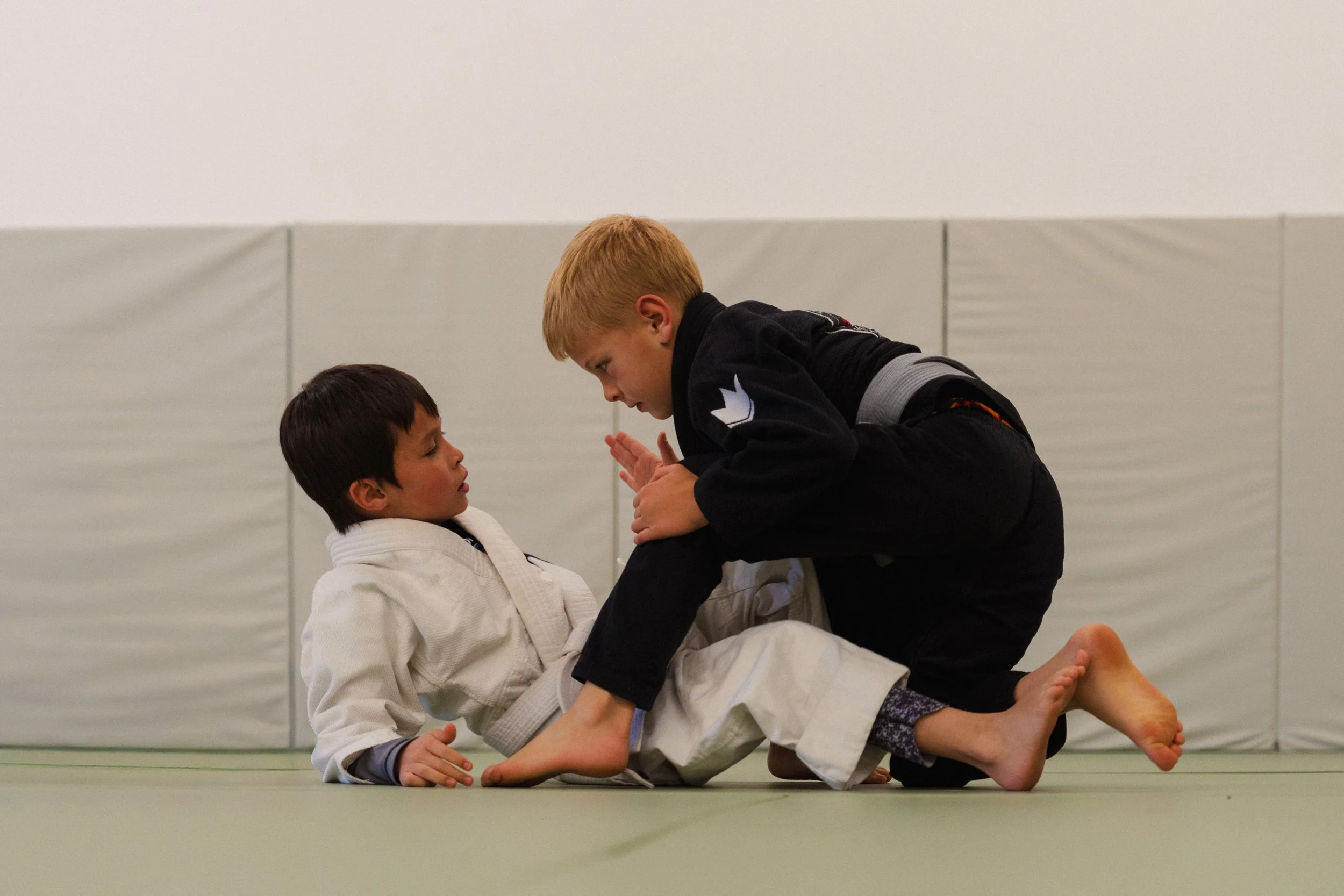 Two children practicing Brazilian Jiu-Jitsu on a mat, one in a white gi and the other in a black gi, engaged in a grappling move.