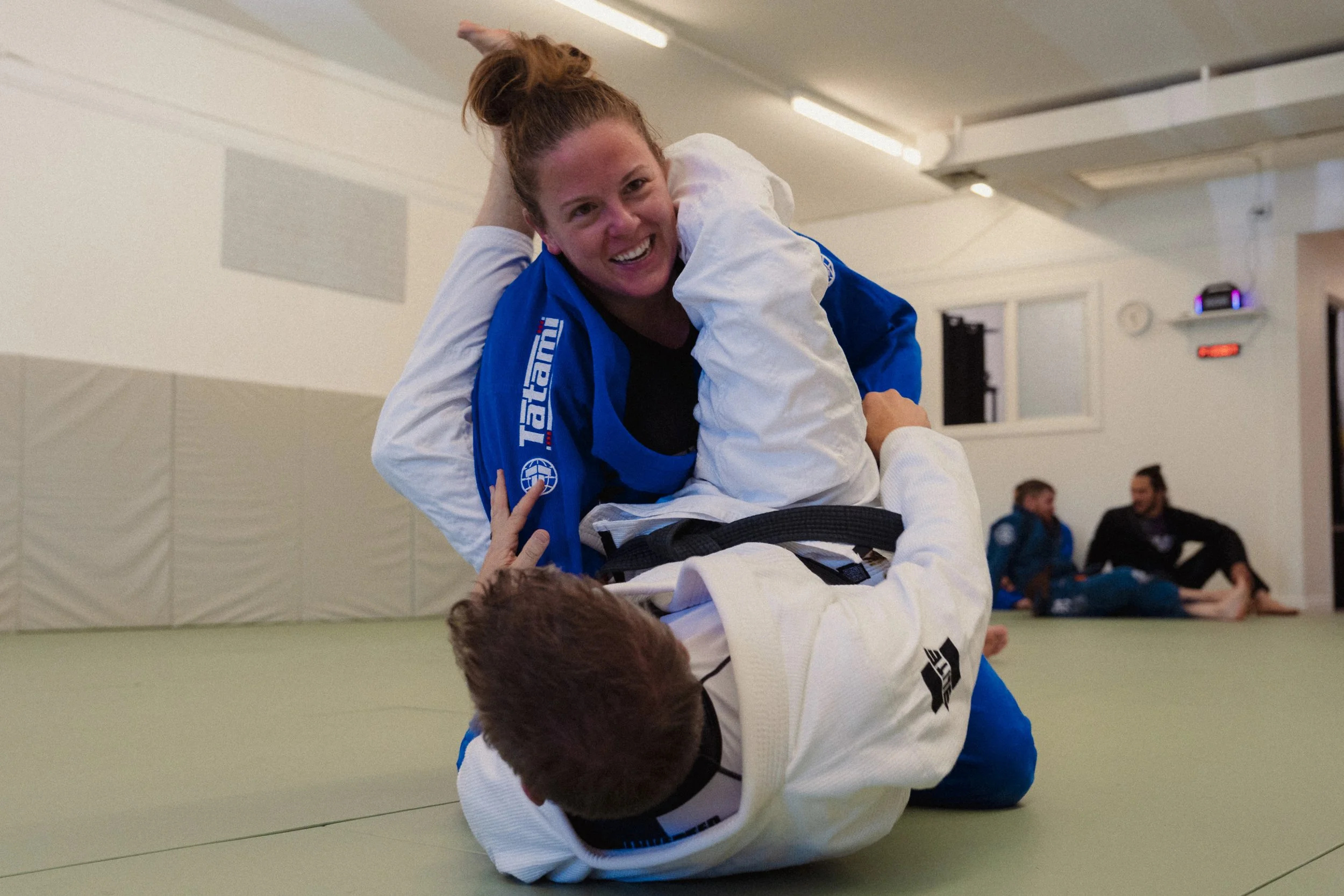 A woman in a blue martial arts uniform practicing Brazilian Jiu-Jitsu with a smile, training on the mat with an opponent during a session in a martial arts gym.