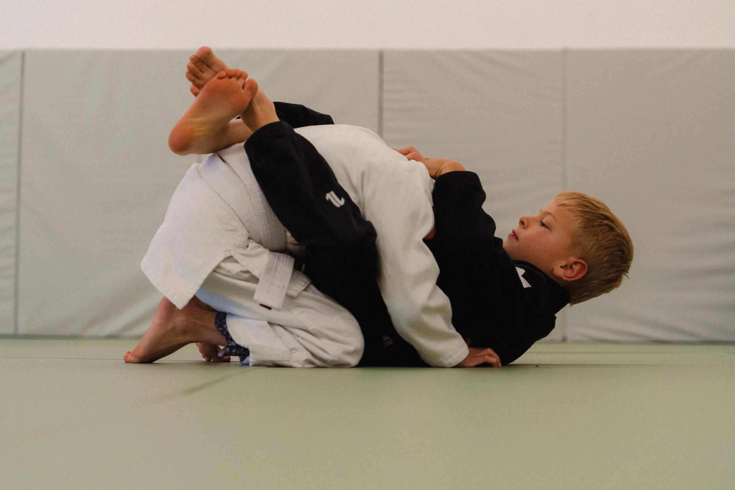 Two children practicing Brazilian Jiu-Jitsu on a mat, wearing gis. One child with a white gi is on their back, while the other child with a black gi is on top, gripping the other child's arm.