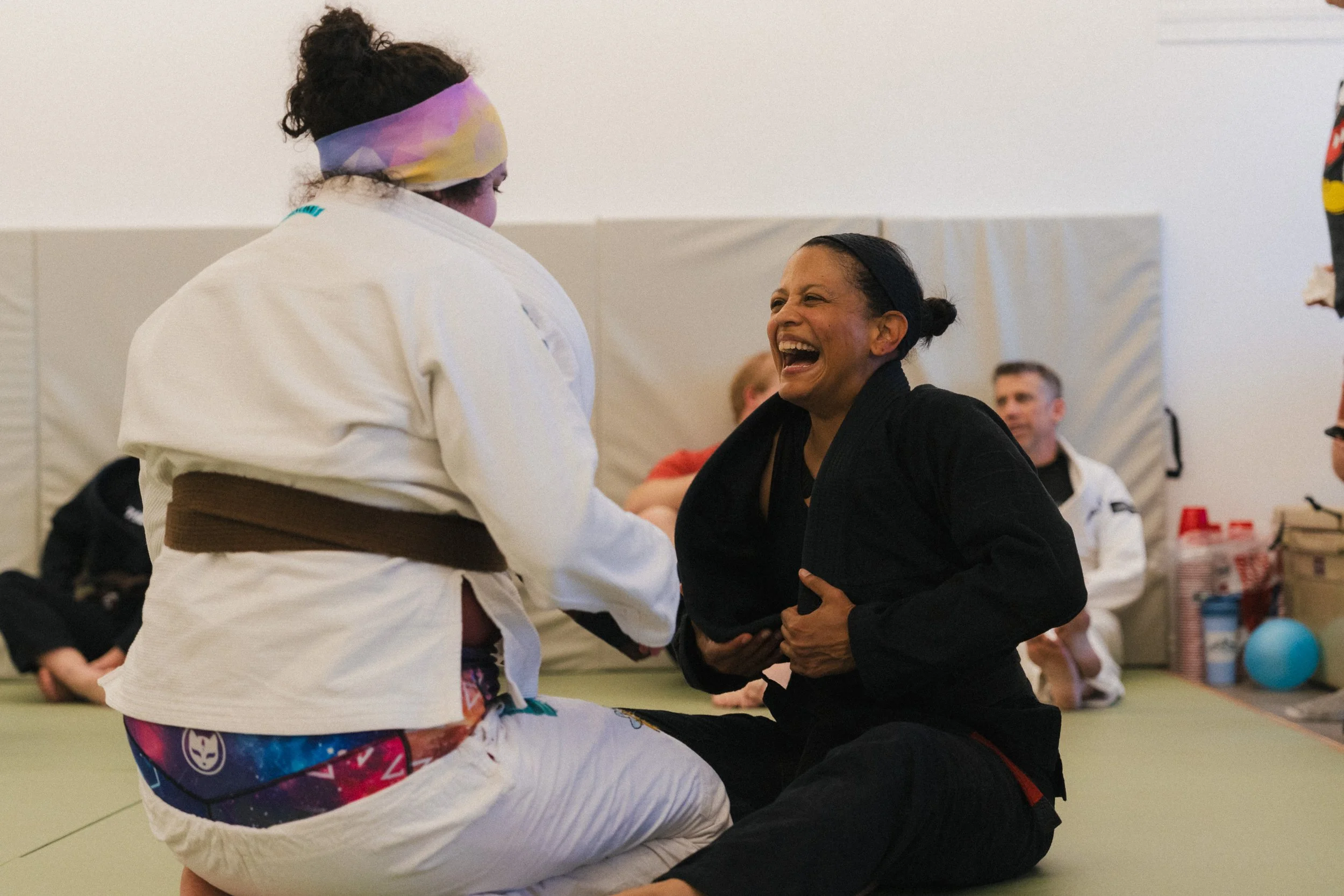 Two women practicing Brazilian Jiu-Jitsu on a green mat in a martial arts gym. One woman is sitting on the floor in a black gi, laughing, while the other woman, wearing a white gi and a colorful headband, appears to be smiling and leaning forward.