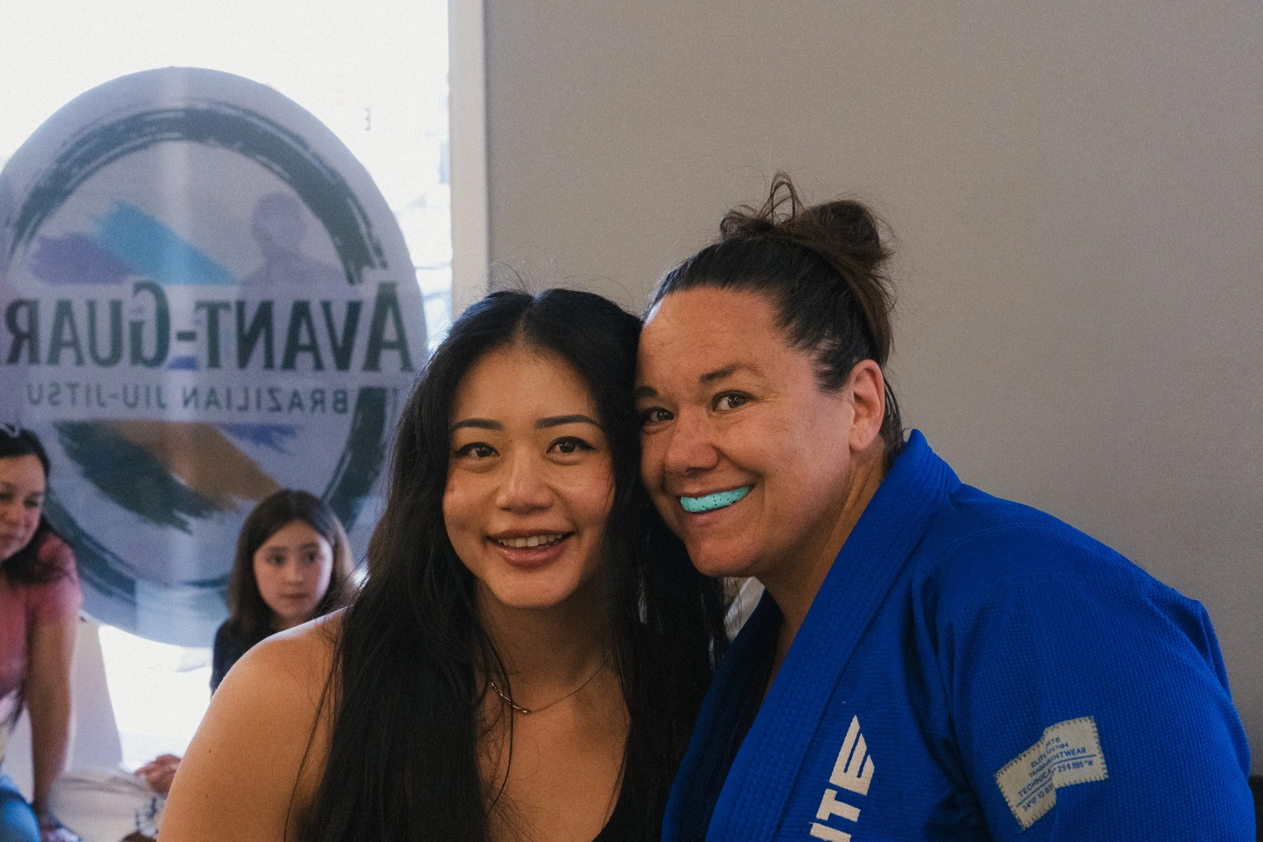 Two women taking a selfie, with a group of people and a sign in the background that says 'VAN-CLUB' and 'BRASILIAN I9-ITU.'