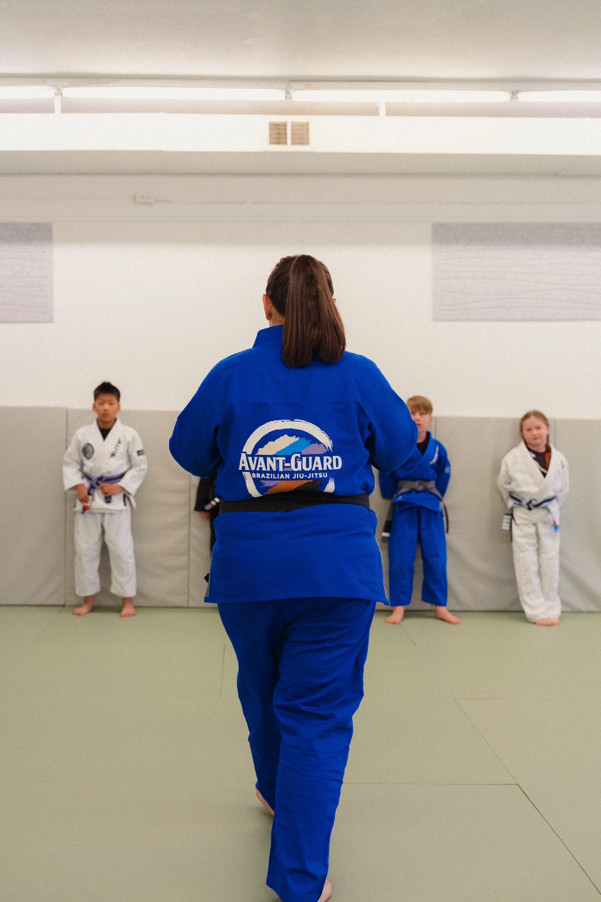 An instructor in a blue gi demonstrating a martial arts move in front of children in martial arts uniforms in a dojo.