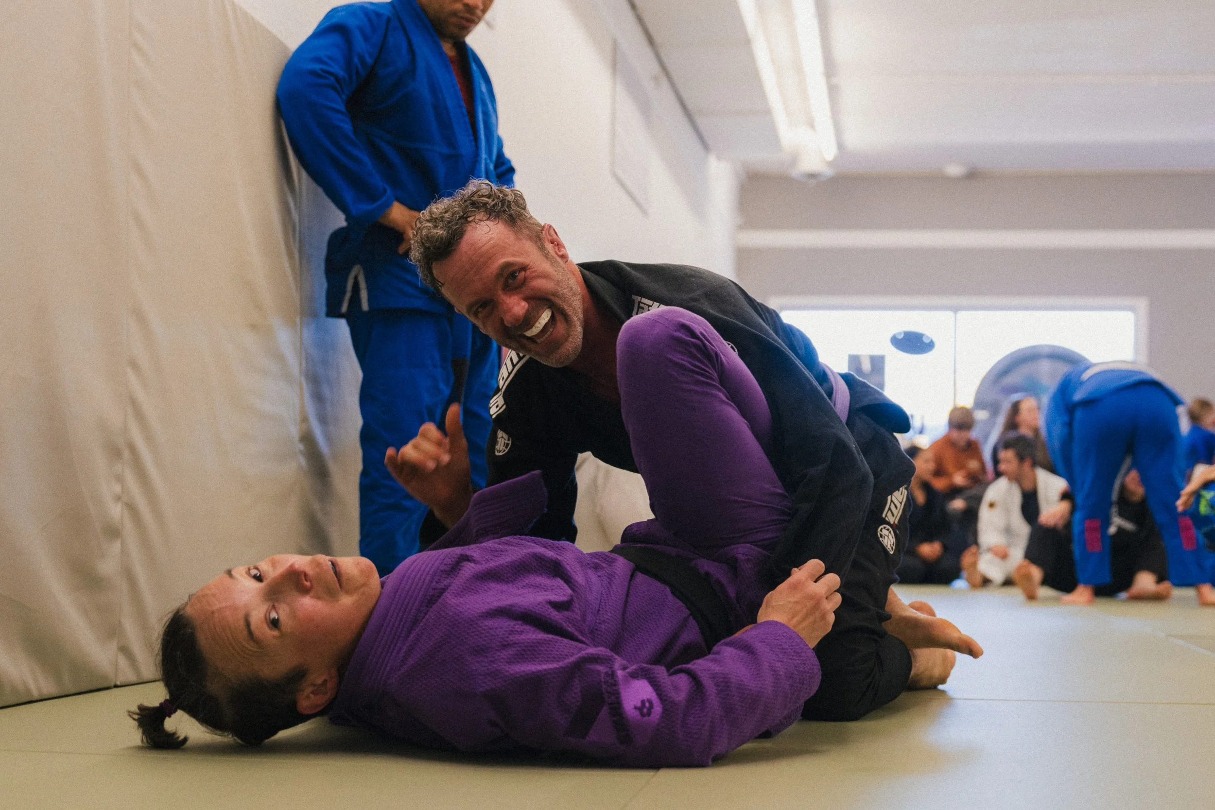 Man and woman practicing Brazilian Jiu-Jitsu on mats, one in a black gi and the other in a purple gi, with a group of people in the background.