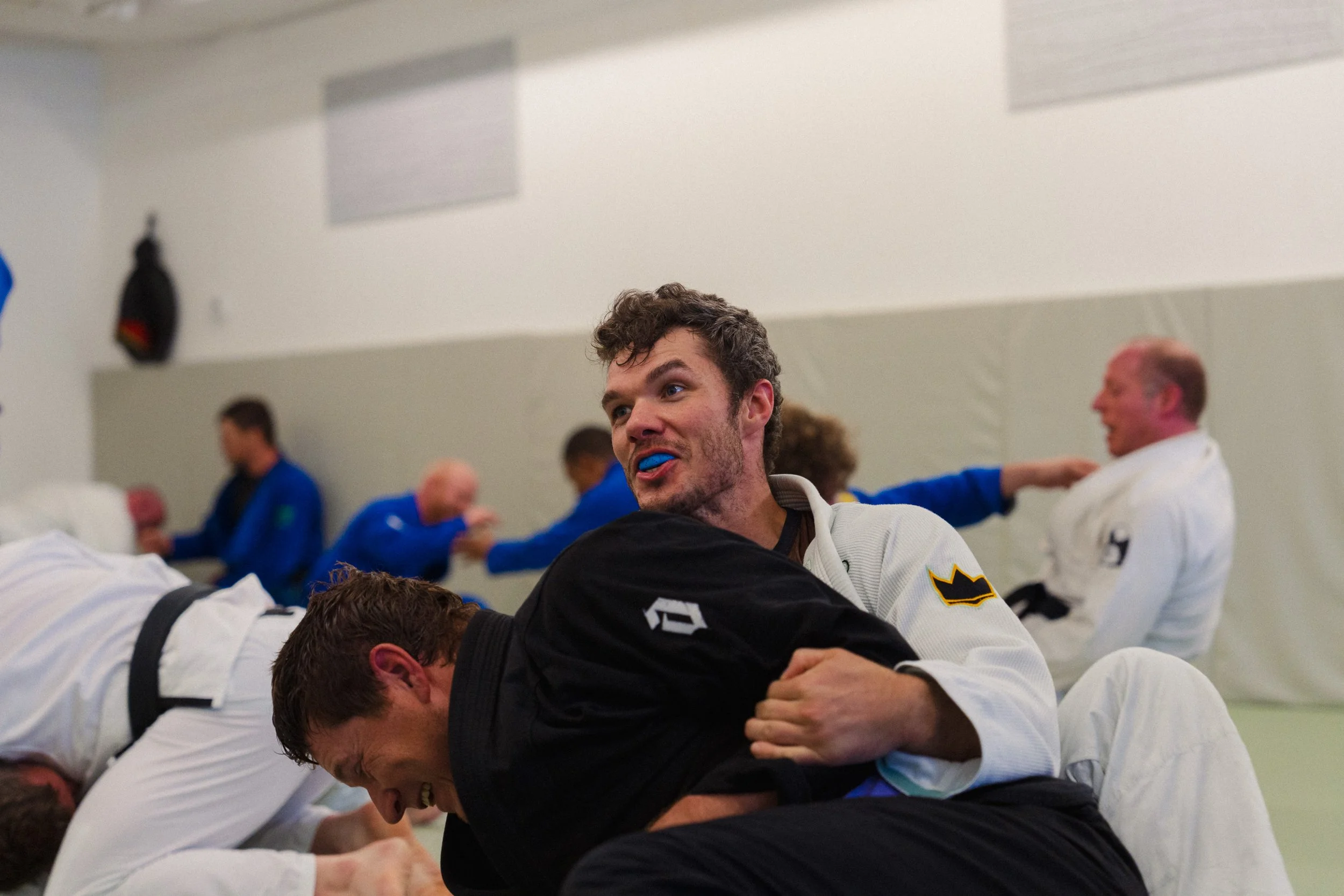 Group of people practicing Brazilian jiu-jitsu, engaged in ground fighting, in a dojo or gym with white walls, some wearing gi uniforms.