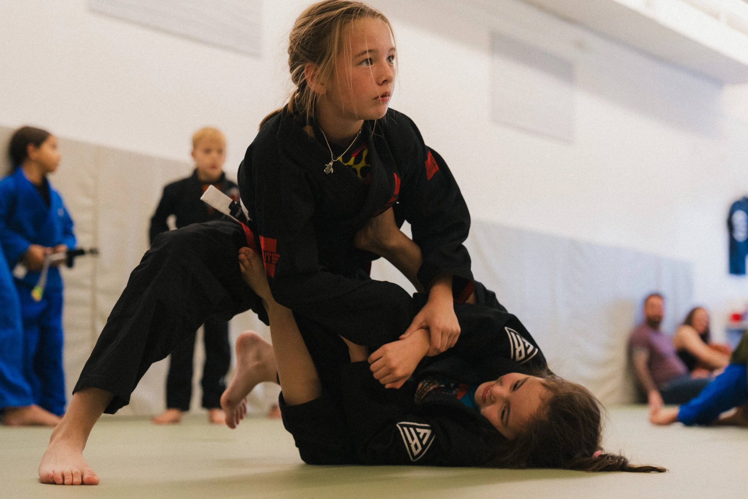 Two young female martial artists practicing Brazilian Jiu-Jitsu in a gym. One girl in a black gi is executing a ground technique while the other girl in a black gi is on the mat, and other children in gi uniforms observe in the background.