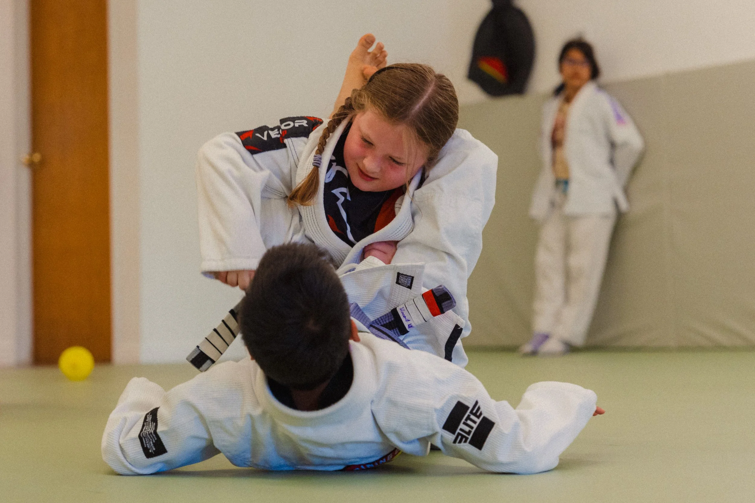 Two young girls in white judo uniforms engaging in a judo match on a mat. One girl is on top, pinning the other girl down, with a woman standing in the background observing. The setting appears to be a martial arts dojo or gym.