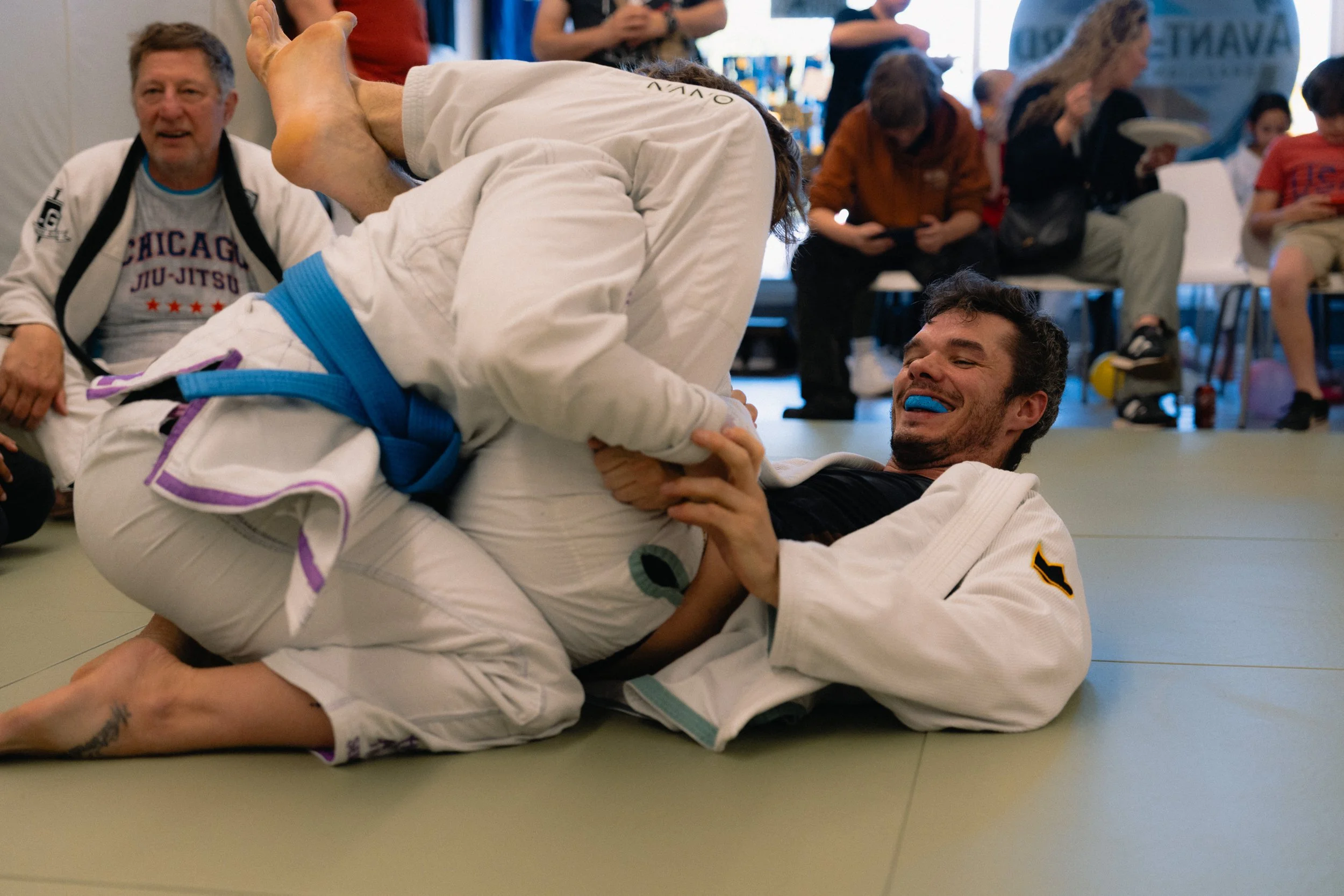 Two men in white Brazilian Jiu-Jitsu gis engaging in a grappling match on a mat, with one man on top and the other on his back. The man on top is holding the other man around the neck and arm during a training session or competition, with several peo
