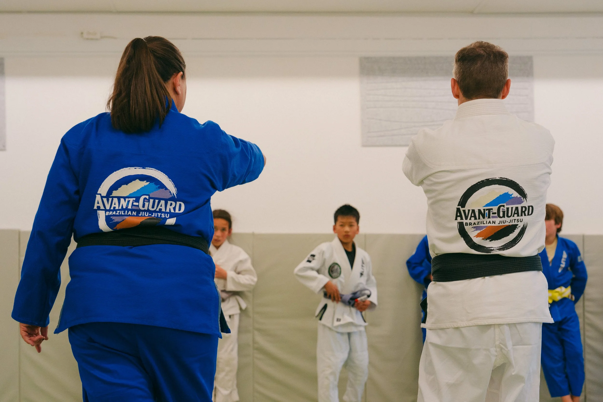 Martial arts instructor and students in a Brazilian Jiu-Jitsu class, wearing gis and belts, in a martial arts dojo.
