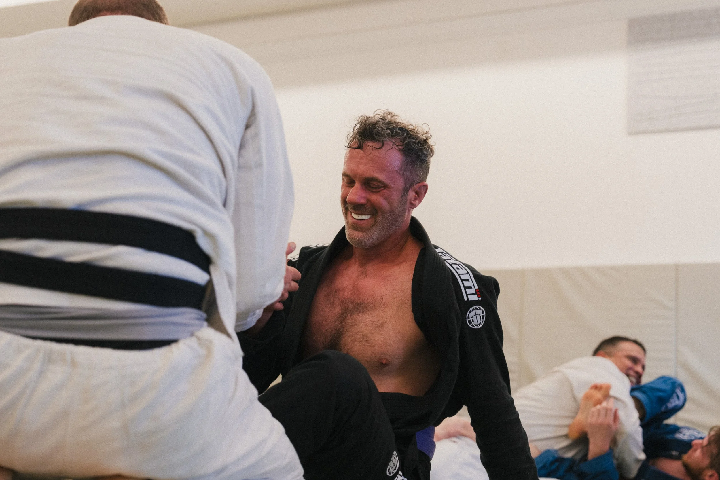 A smiling man with curly hair and a beard is wearing a black martial arts gi with patches and is smiling while standing in a dojo. Other people are also present, some wearing gis, and they are sitting or kneeling on the mat, possibly during a martial