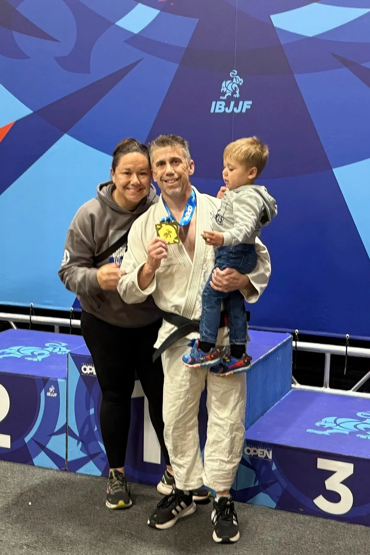 A man wearing a white judo gi holding a gold medal, standing beside a young boy in a gray hoodie and jeans; a woman in a gray hoodie is standing next to them. They are on a podium with a blue backdrop bearing a logo with a lion and the letters "IBJJF" and a number 3, indicating third place at a judo or Brazilian jiu-jitsu competition.