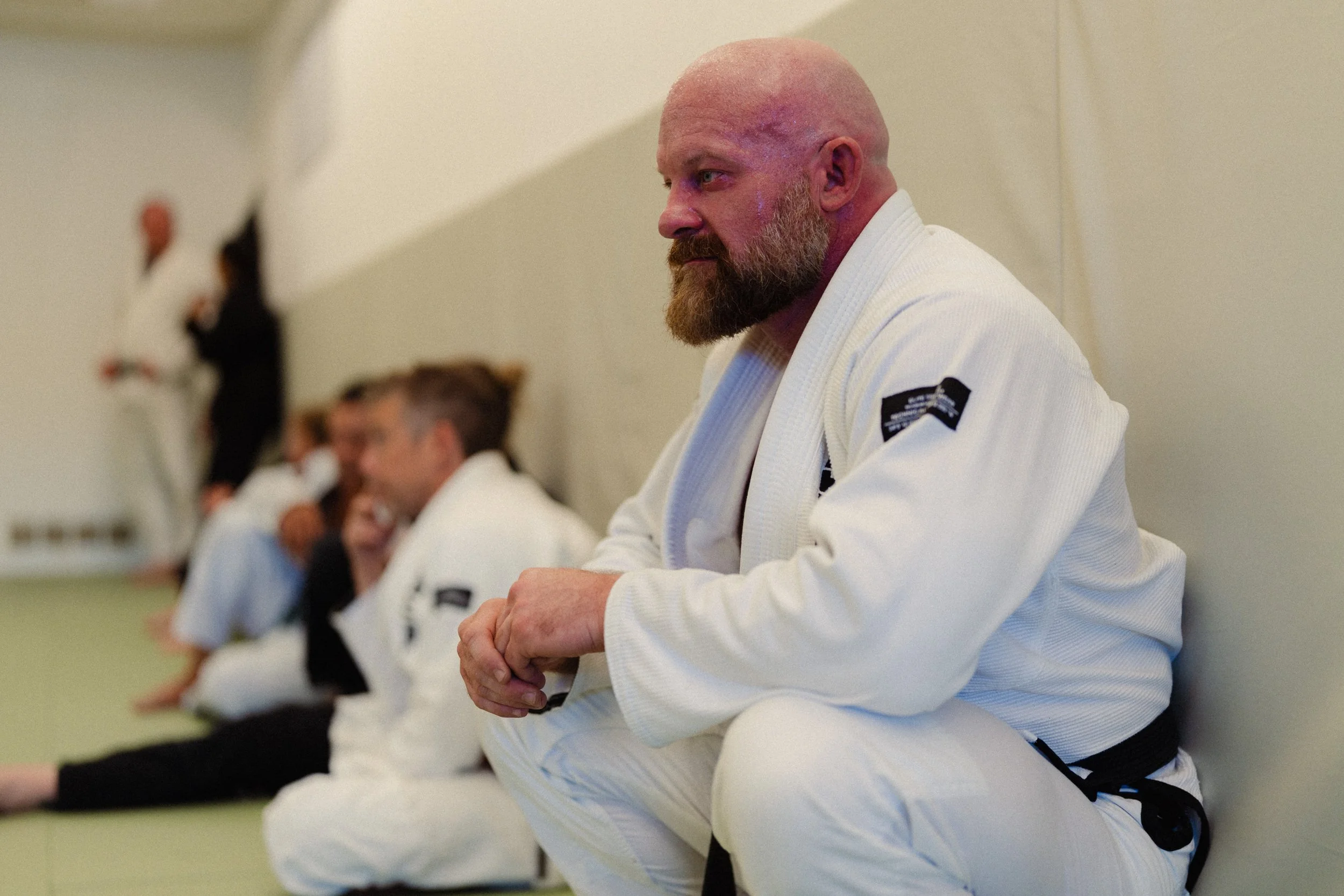 A group of men in white judo uniforms sitting cross-legged on the floor, with their hands clasped, in an academy setting, preparing for or engaged in a martial arts class.