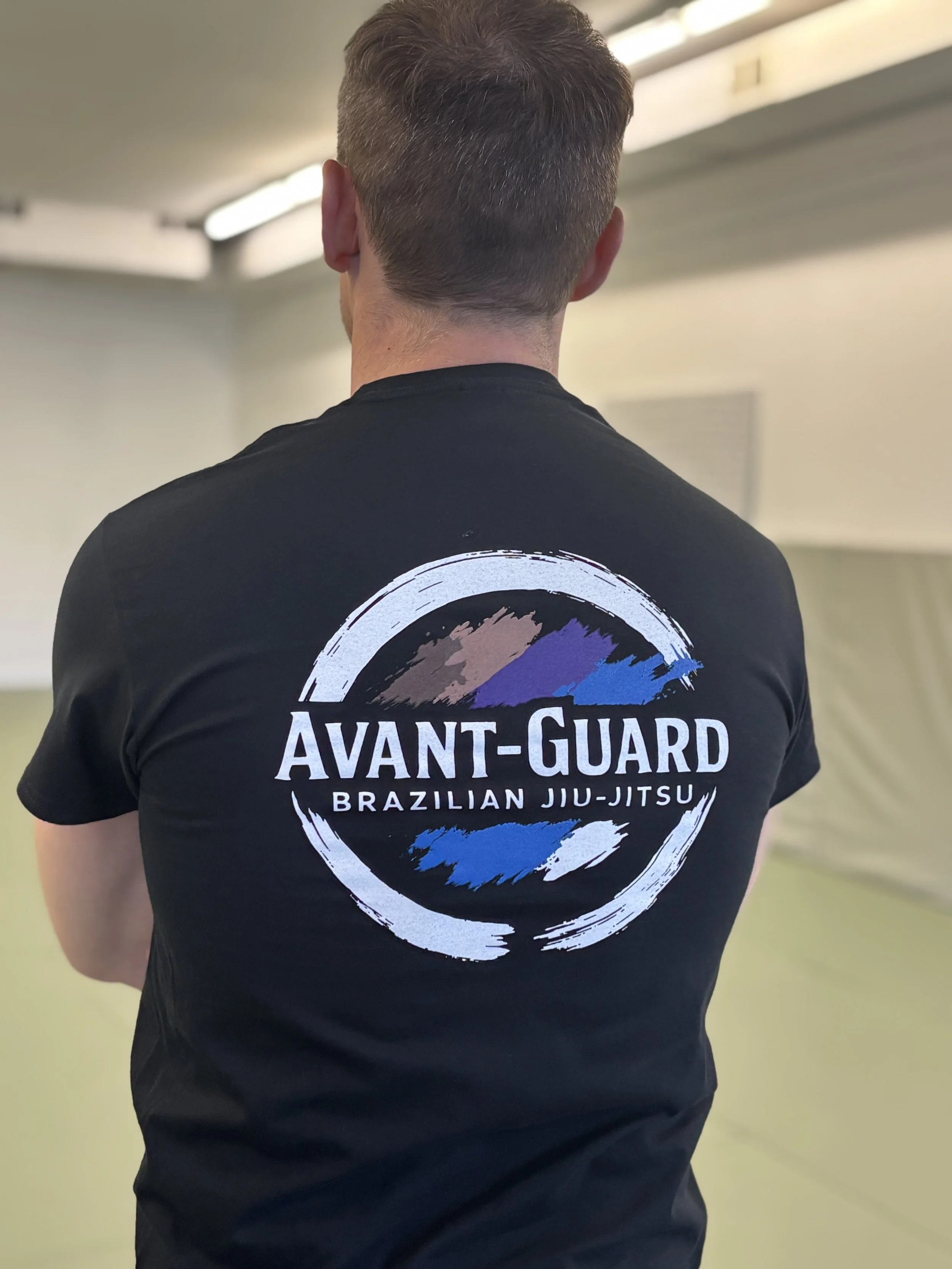 Back of a person wearing a black T-shirt with the logo 'Avant-Guard Brazilian Jiu-Jitsu' and a colorful graphic on the back, standing in an indoor sports facility.